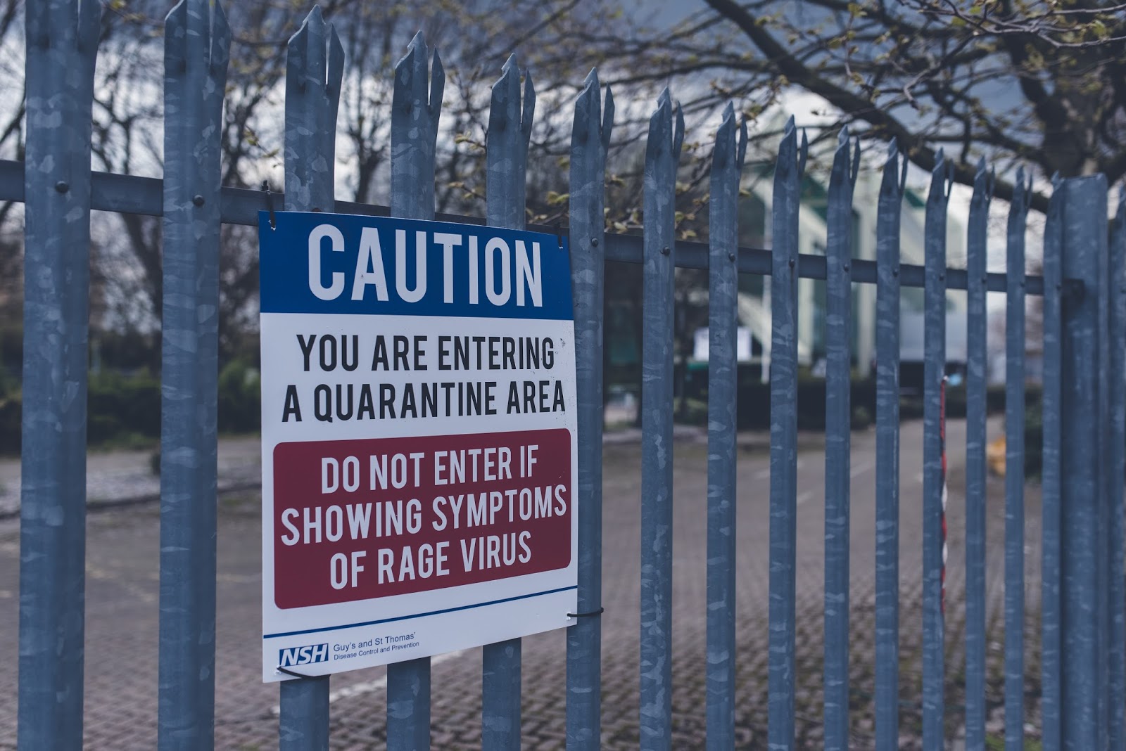 A metal fence with a sign that reads, "CAUTION: YOU ARE ENTERING A QUARANTINE AREA. DO NOT ENTER IF SHOWING SYMPTOMS OF RAGE VIRUS." The sign includes a logo for NSH. In the background, blurred trees and a path are visible. The photo is a great way to share a sneak peak of what the event was like for a Secret Cinema review.