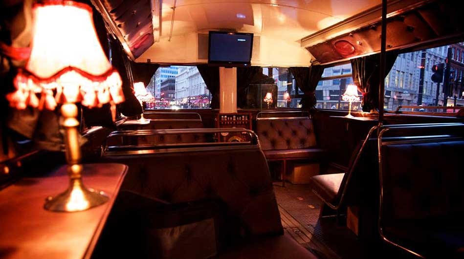This image shows the interior of a vintage-style bus turned into a cozy dining area. It features tufted leather seating, small tables with ornate lamps, and a view of a busy city street through the windows. The atmosphere is warm and inviting, perfect for reminiscing about Halloween 2016.