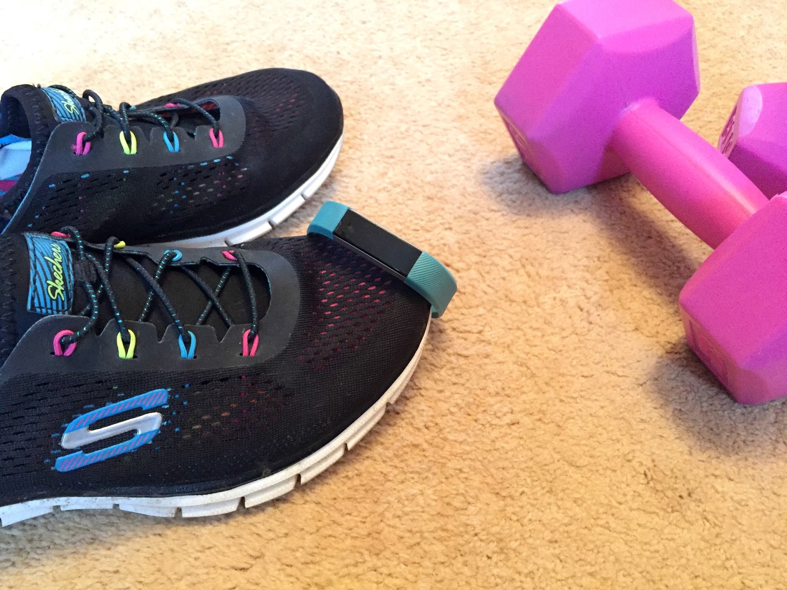 A pair of black trainers with a turquoise Fitbit resting on top. Beside them are some small pink weights.