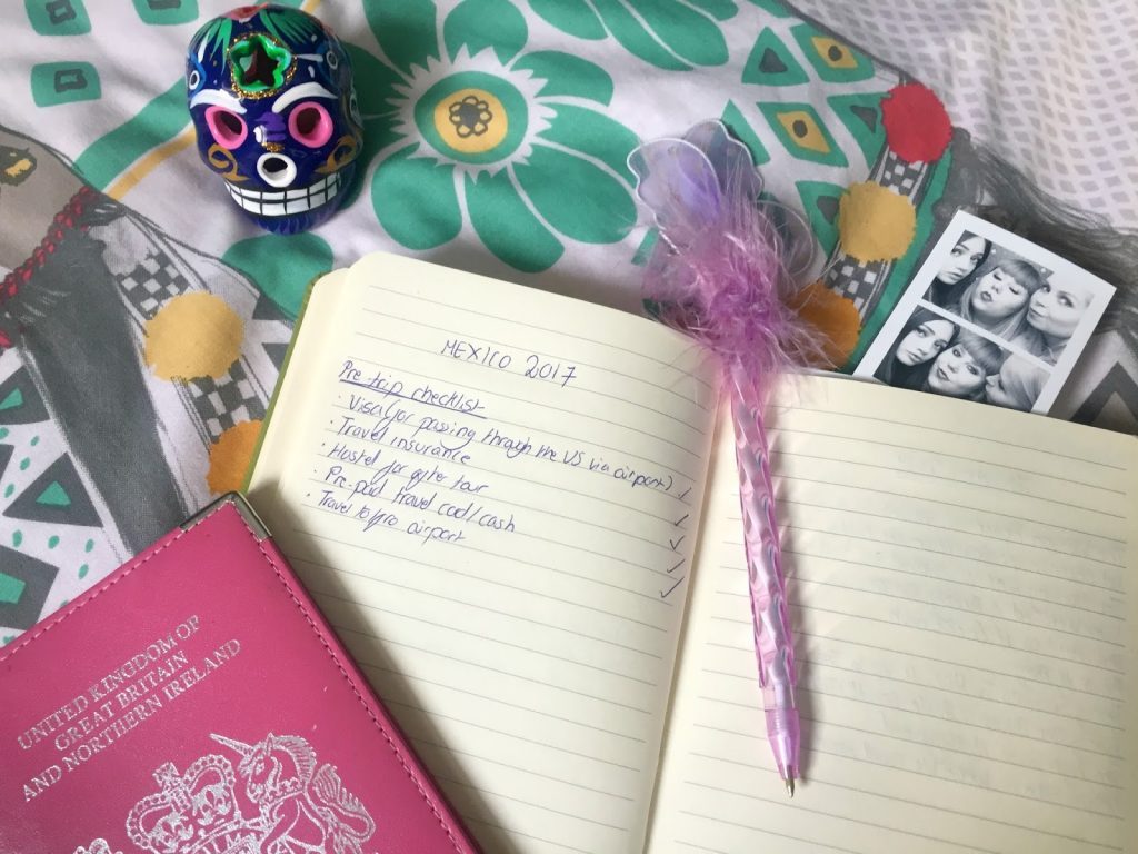 An open journal with "Mexico 2017" and a travel checklist, surrounded by a feather pen, a small passport with the United Kingdom cover, a colorful decorated skull, a patterned background, and a black-and-white photobooth strip featuring two people celebrating their adventurous career.