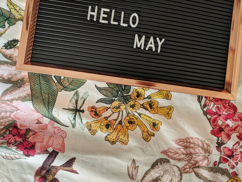 A wooden-framed black letter board with the words "HELLO MAY" in white letters lies on a floral-printed fabric. The fabric showcases various flowers and leaves in shades of pink, yellow, green, and brown, complemented by an image of a dragonfly.