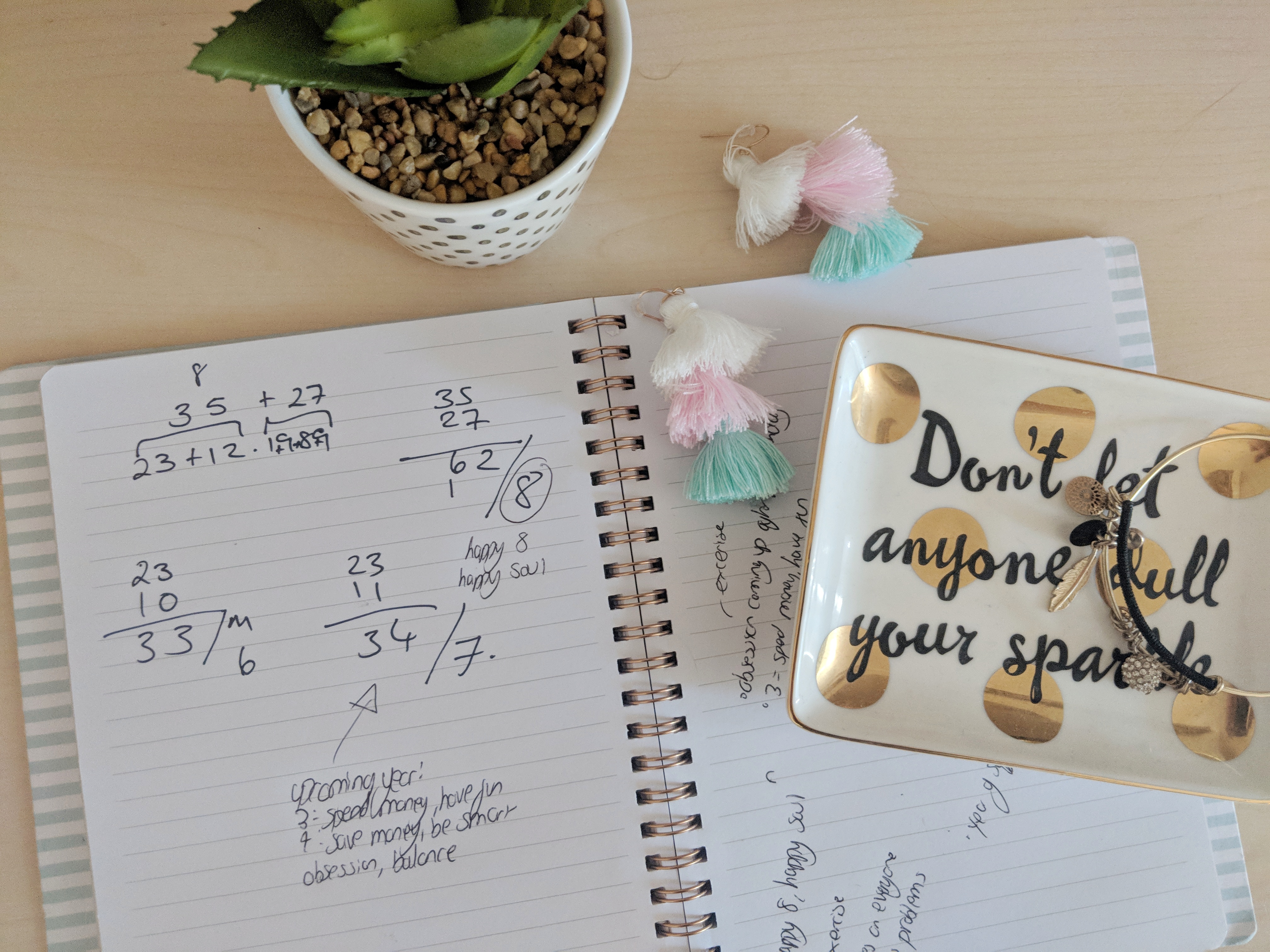 An open notebook with doodles and math problems, perhaps hinting at some hidden numerology. Beside it are a potted succulent, a small decorative dish with "Don't let anyone dull your sparkle" written on it, and some pastel-colored tassel keychains. The scene is set on a light-colored desk.