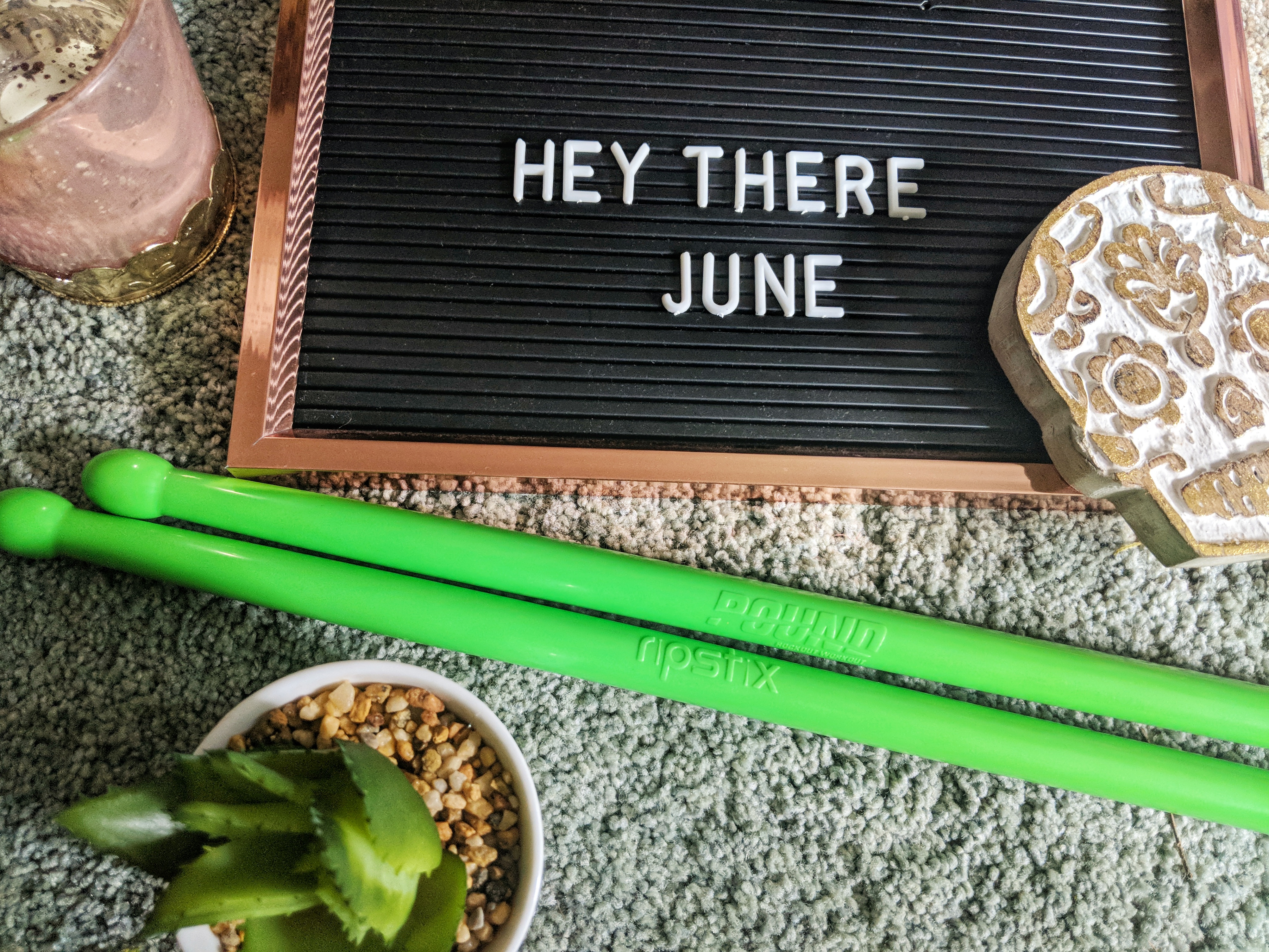A decorative board with the message "HEY THERE JUNE" is placed on a textured surface, surrounded by various items, including bright green Ripstix drumsticks, a small potted succulent, a patterned coaster, and a glass of smoothie.