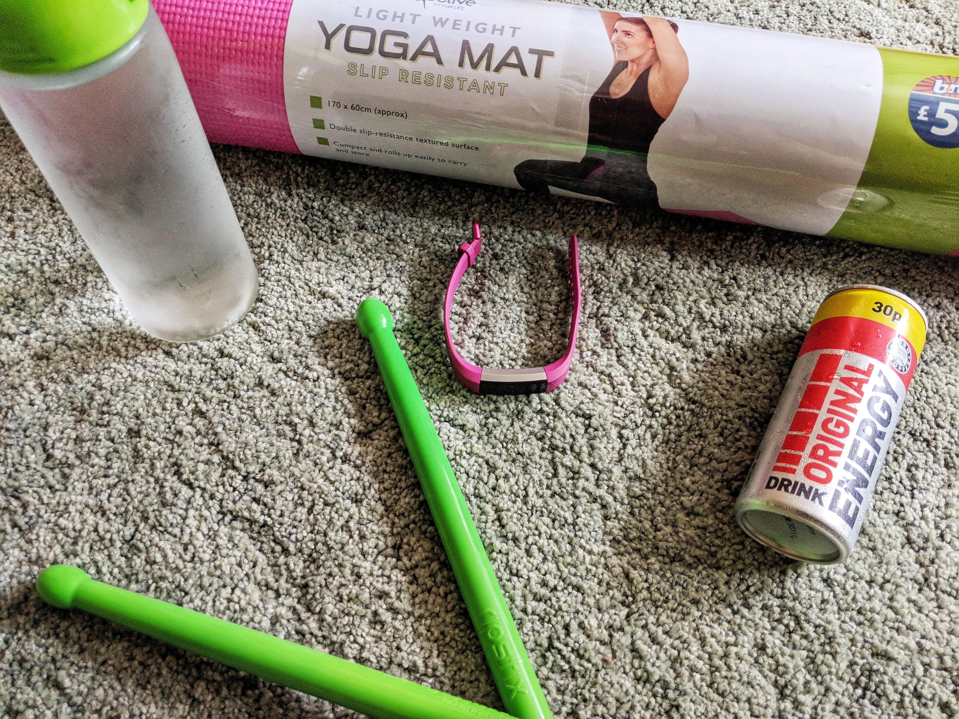 A rolled pink yoga mat, a water bottle, a fitness tracker with a pink strap, two green drumsticks for home workouts, and a can of energy drink are arranged on a gray carpet.