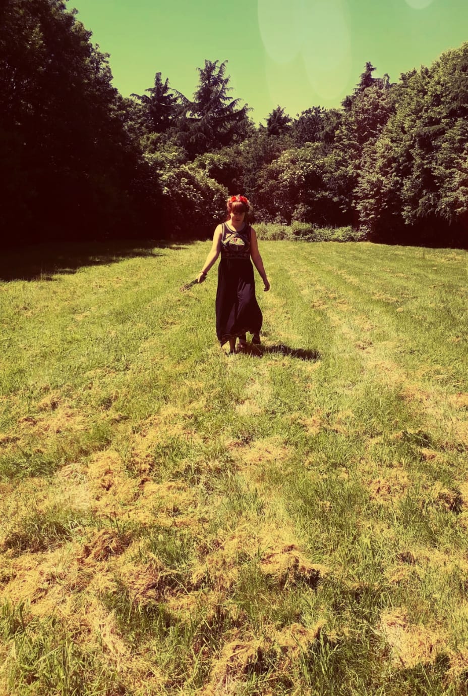 Louise is walking through a field, wearing a long flowing black dress with boho-style patterning on the top. She is holding some daffodils in her hand and wearing a flower crown.