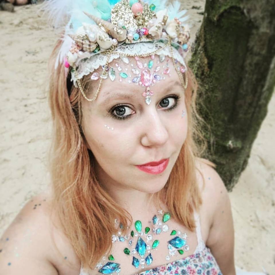 Louise wears an elaborate headpiece adorned with shells, pearls, and feathers. Their face and upper chest are decorated with colorful gemstones.