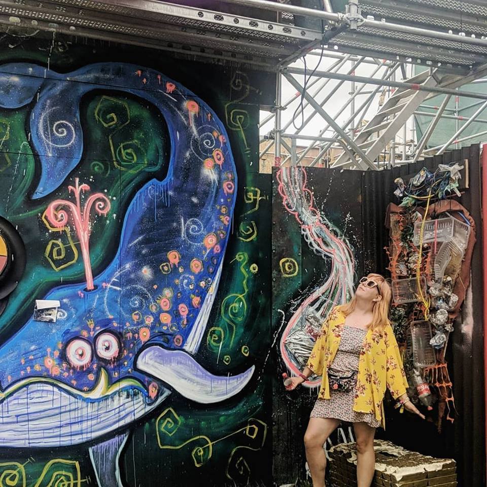 Louise with long blonde hair, wearing a yellow floral jacket and sunglasses, stands smiling in front of a vibrant mural featuring a whimsical sea creature and colorful abstract designs. She appears to be in an industrial setting with exposed metal scaffolding above, reminiscent of the lively backdrop one might find at Boomtown Fair.