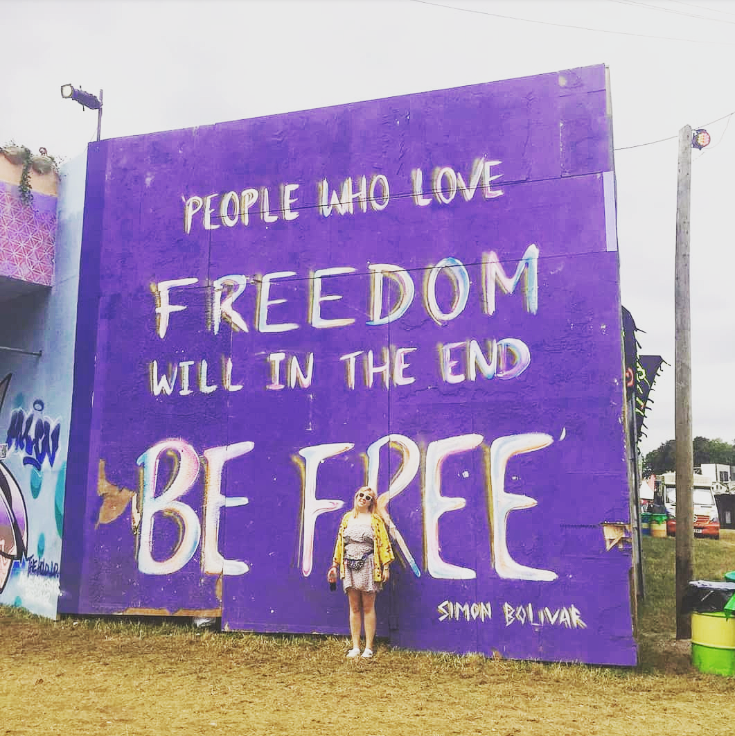 Louise is standing in front of a huge purple sign at Boomtown Music Festival. The sign reads 'people who love freedom will in the end be free' by Simon Bolivar. She is wearing a short pale pink patterned dress with a bright yellow jacket.