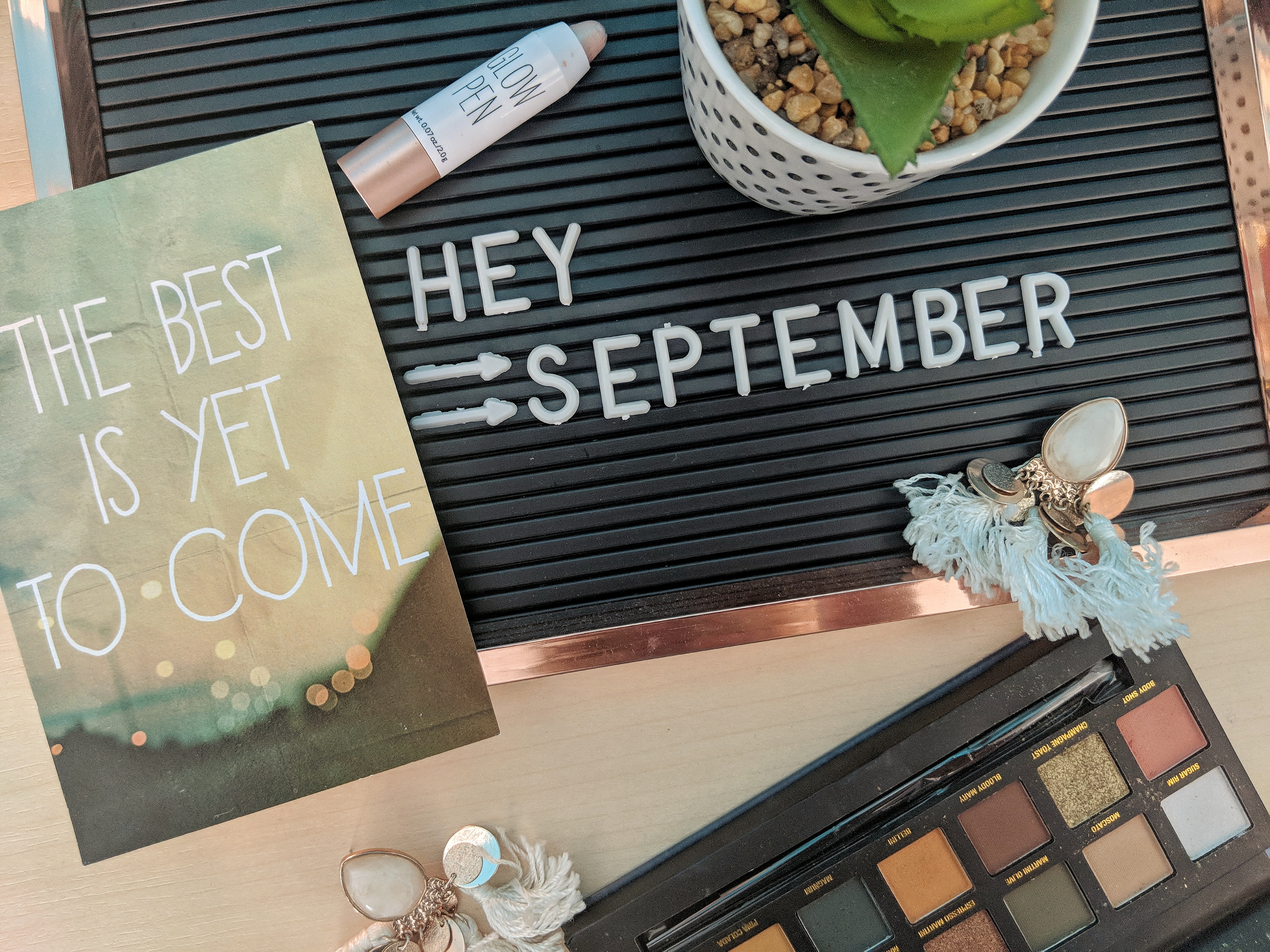 A flat lay photo captures a letter board with the text "HEY SEPTEMBER," a card reading "THE BEST IS YET TO COME," a bottle of glow pen, a small potted plant, a pair of earrings, and an open eyeshadow palette with various colors.
