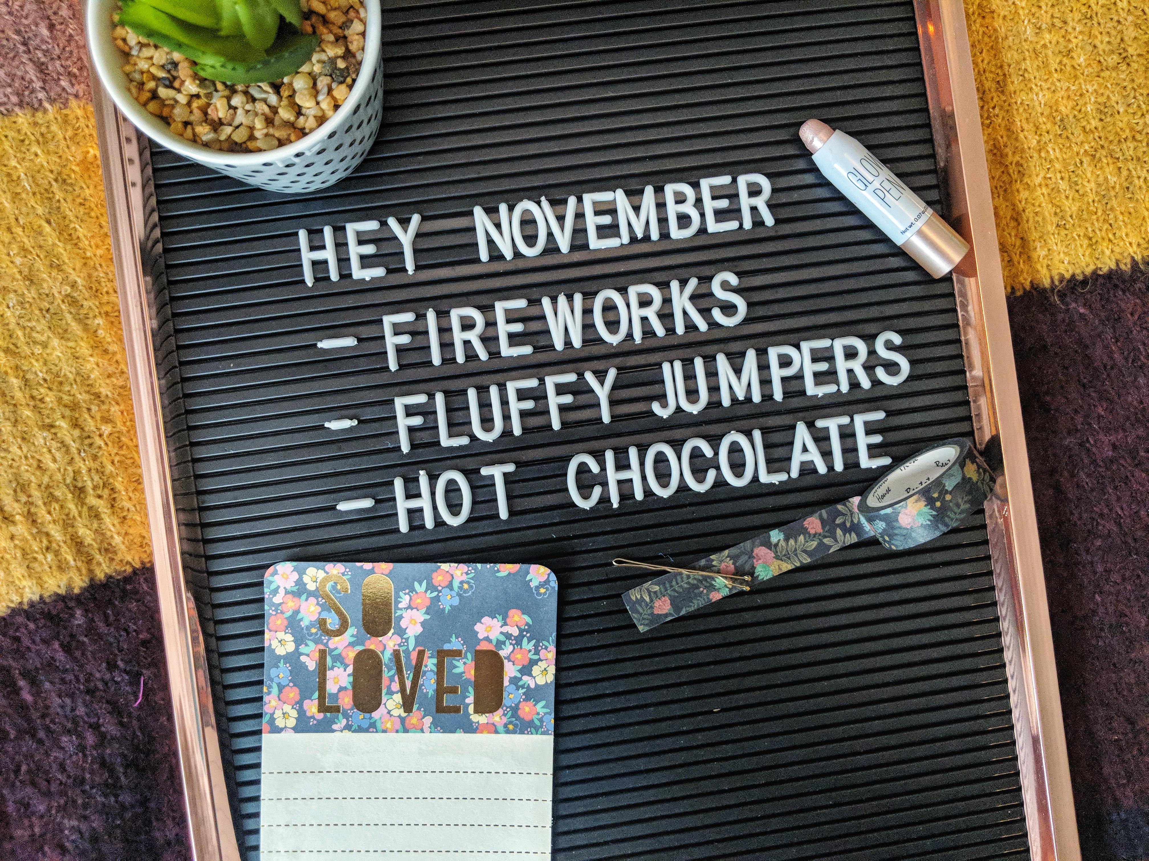A letter board reads "Hey November - Fireworks - Fluffy Jumpers - Hot Chocolate." Surrounding the board are a small plant, a highlighter, a roll of decorative tape, a polka-dot notebook with "So Loved" on the cover, and a gold bobby pin.