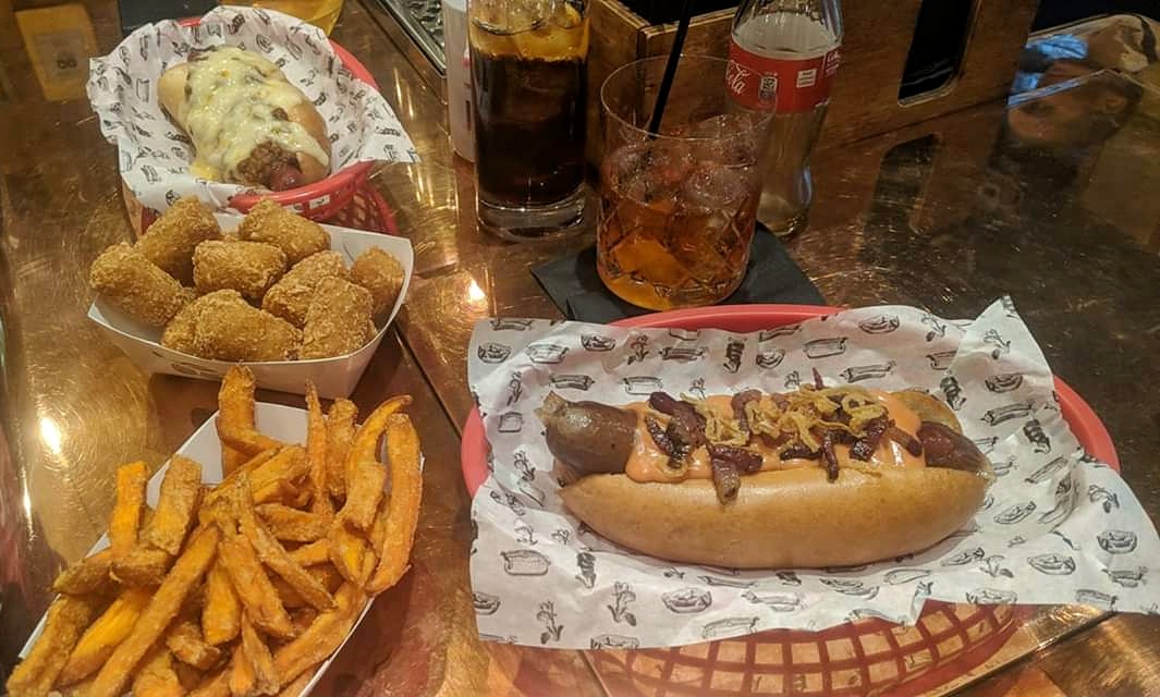 A meal on a London table includes two hot dogs in paper-lined baskets, one topped with caramelized onions and the other with melted cheese. Sides of sweet potato fries and breaded potato bites are also present. Drinks include a soda in a glass and a bottle of Coca-Cola.