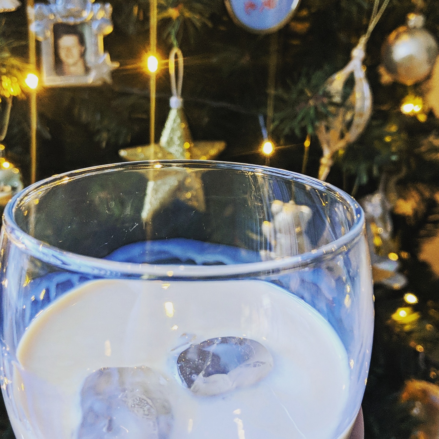A close-up of a glass of creamy white drink with ice cubes, held in front of a decorated Christmas tree. The tree is adorned with golden star ornaments, lights, and a framed photo ornament, conjuring the festive holiday spirit typical of December.