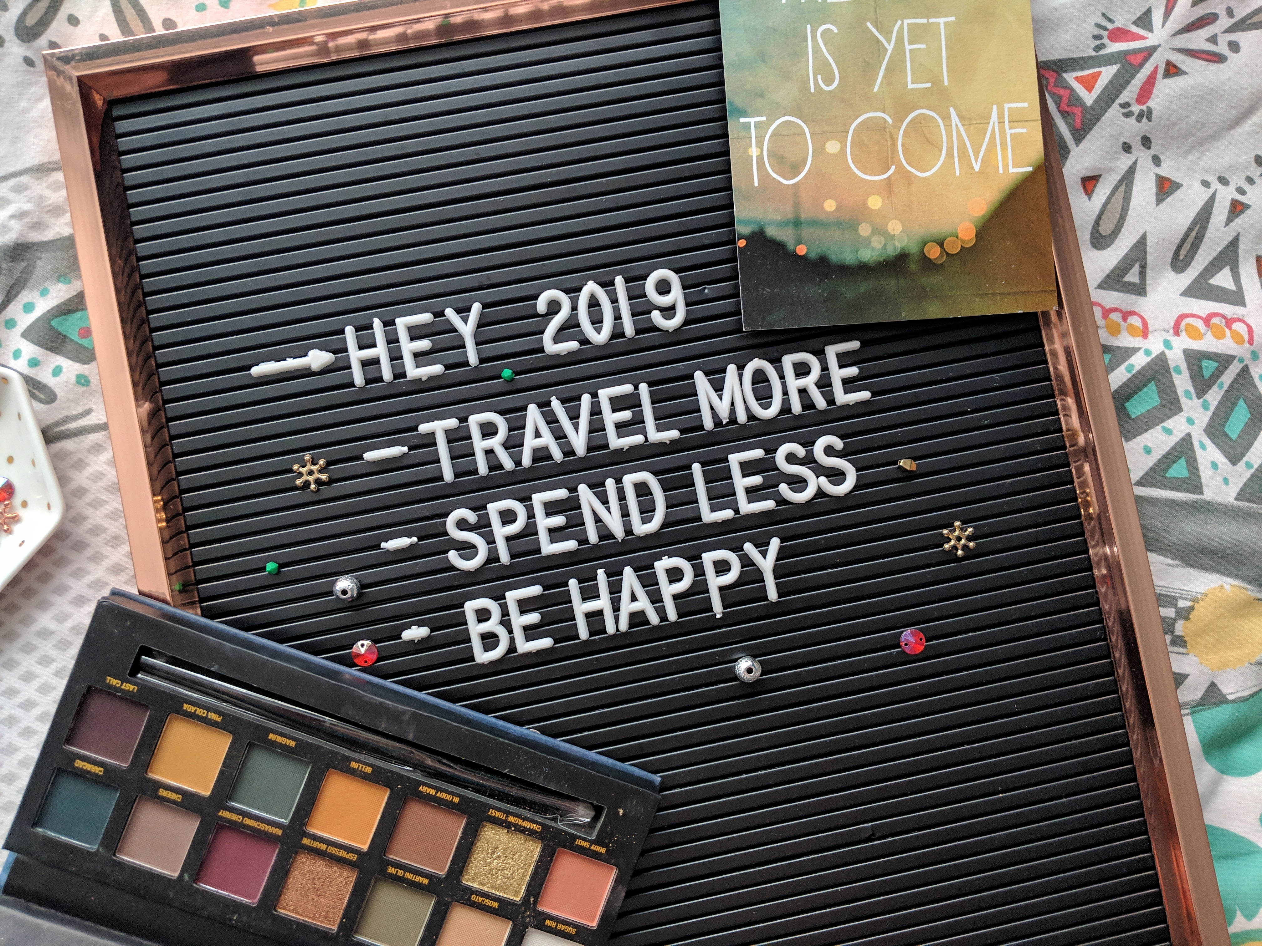 A letter board displays the message "HEY 2019 - TRAVEL MORE, SPEND LESS, BE HAPPY." Beside the board is an eyeshadow palette and a card with the words "IS YET TO COME" against a colorful January backdrop. The arrangement is atop a patterned fabric.