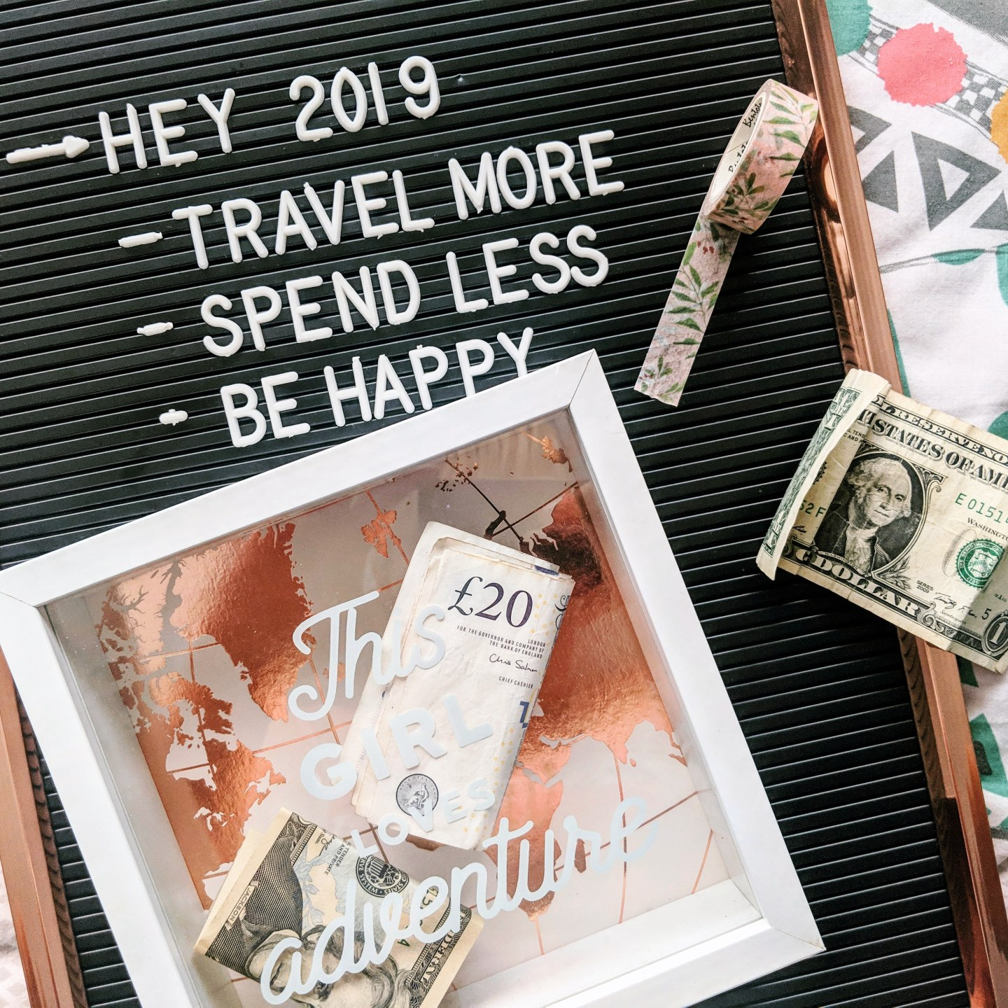 A money travel box which says 'this girl loves adventure' on the front in cursive white writing. There is money inside. The box is laying on a letter board which lists Louise's 2019 goals, including to travel more.