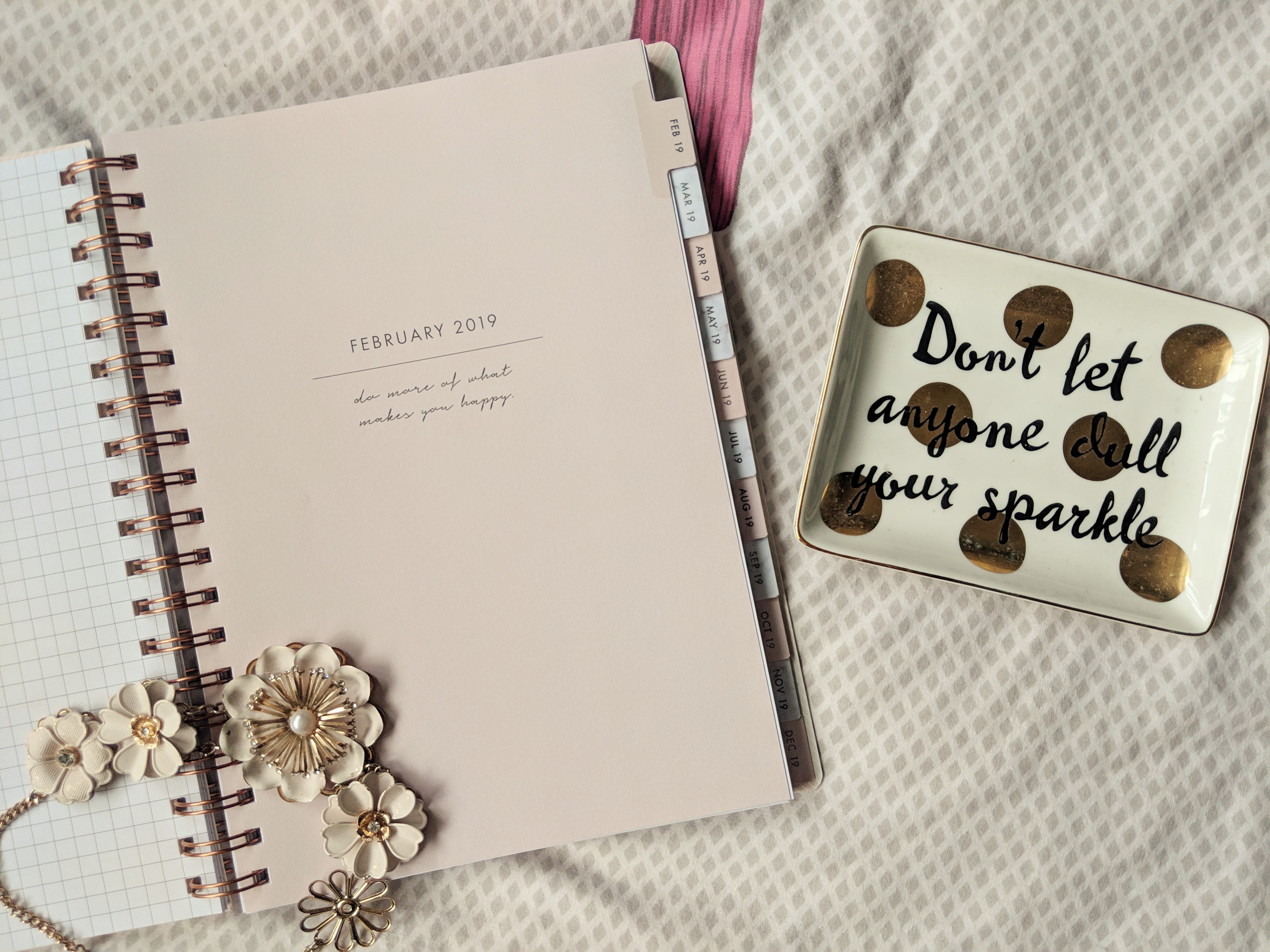 A flat lay of a planner and jewellery in a matching colour lying on top. There is also a trinket dish to the side that reads 'don't let anyone dull your sparkle' in cursive writing.
