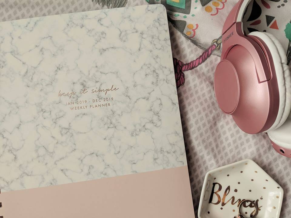 A marble-patterned weekly planner, titled "Keep it Simple," sits next to a pair of pink headphones. The planner dates from January 2019 to April 2019. Nearby is a small dish with the word “Bliss” on it. The background includes colorful fabric with various patterns.