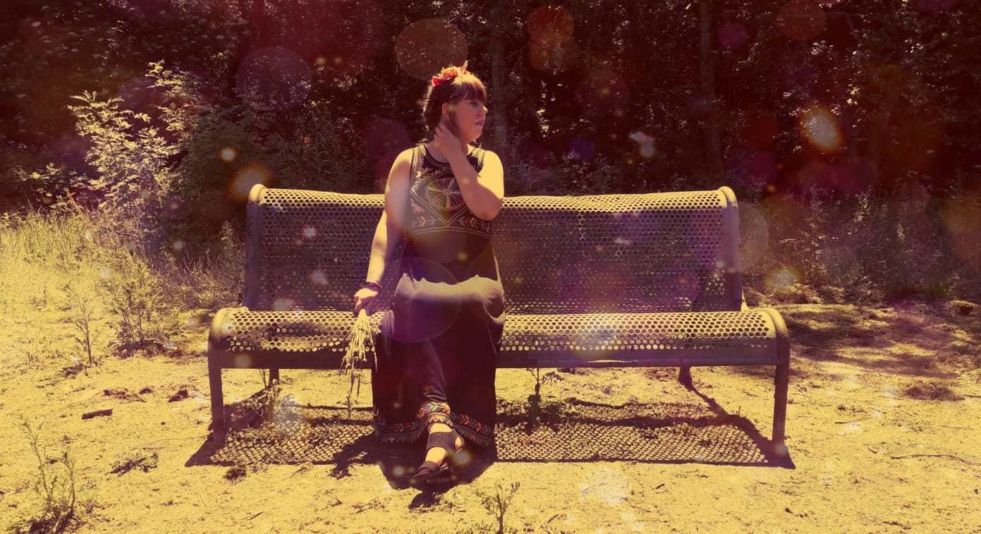 Louise is sitting on a bench, wearing a long flowing black dress with boho-style patterning on the top. She is holding some daffodils in her hand and wearing a flower crown.