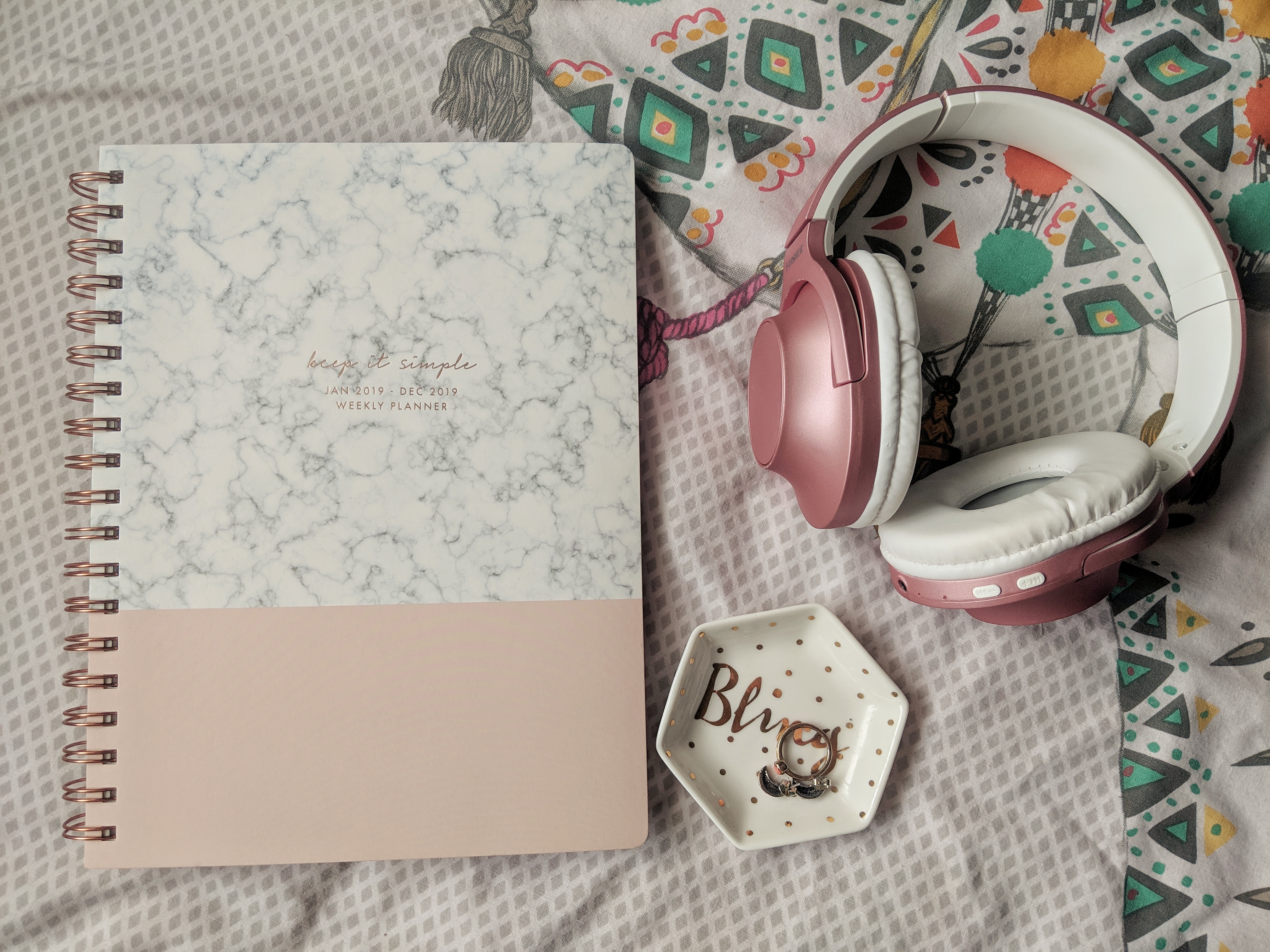 An open journal with a marbled and pink cover titled "Weekly Planner: March Edition" lies on a patterned bedsheet next to folded pink and white headphones. A hexagonal dish with the word "Hello" and a ring inside it rests nearby.