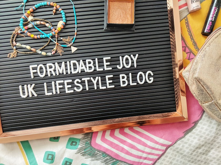 A flat lay photo featuring a letter board that reads "Formidable Joy - UK Lifestyle Blog". Surrounding the board are colorful beaded necklaces, a small lidded box, a makeup brush, printed fabric, and a mesh pouch. The setup captures the vibrant essence of May.