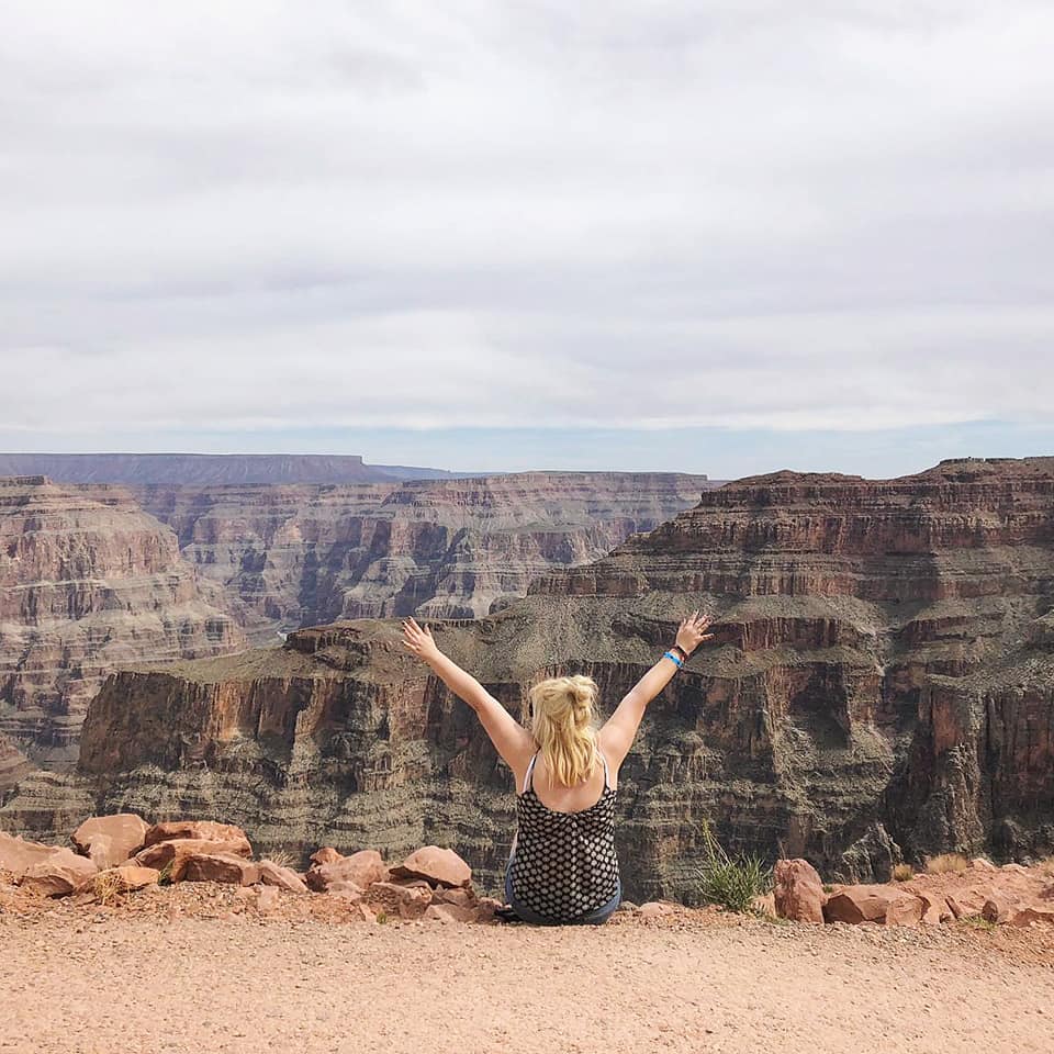 Louise with blonde hair, wearing a black and white polka dot dress, sits at the edge of a cliff with her arms raised in joy, facing the expansive view of the Grand Canyon in the background. The sky is partially cloudy. This moment captures an unforgettable entry in her USA Travel Diary.