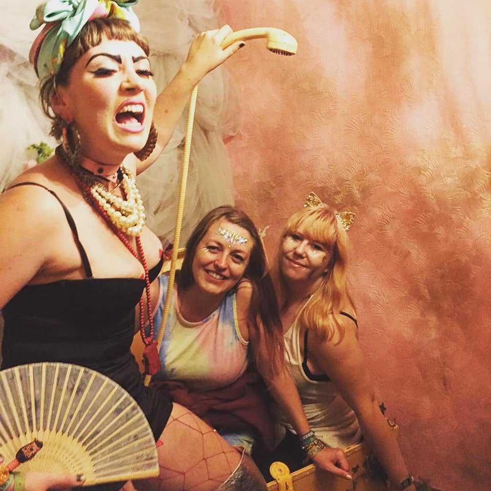Louise and Briony are posing next to an actor at Boomtown Fair. The actor is eccentrically holding a wired telephone above their heads as they pose in a bath tub.