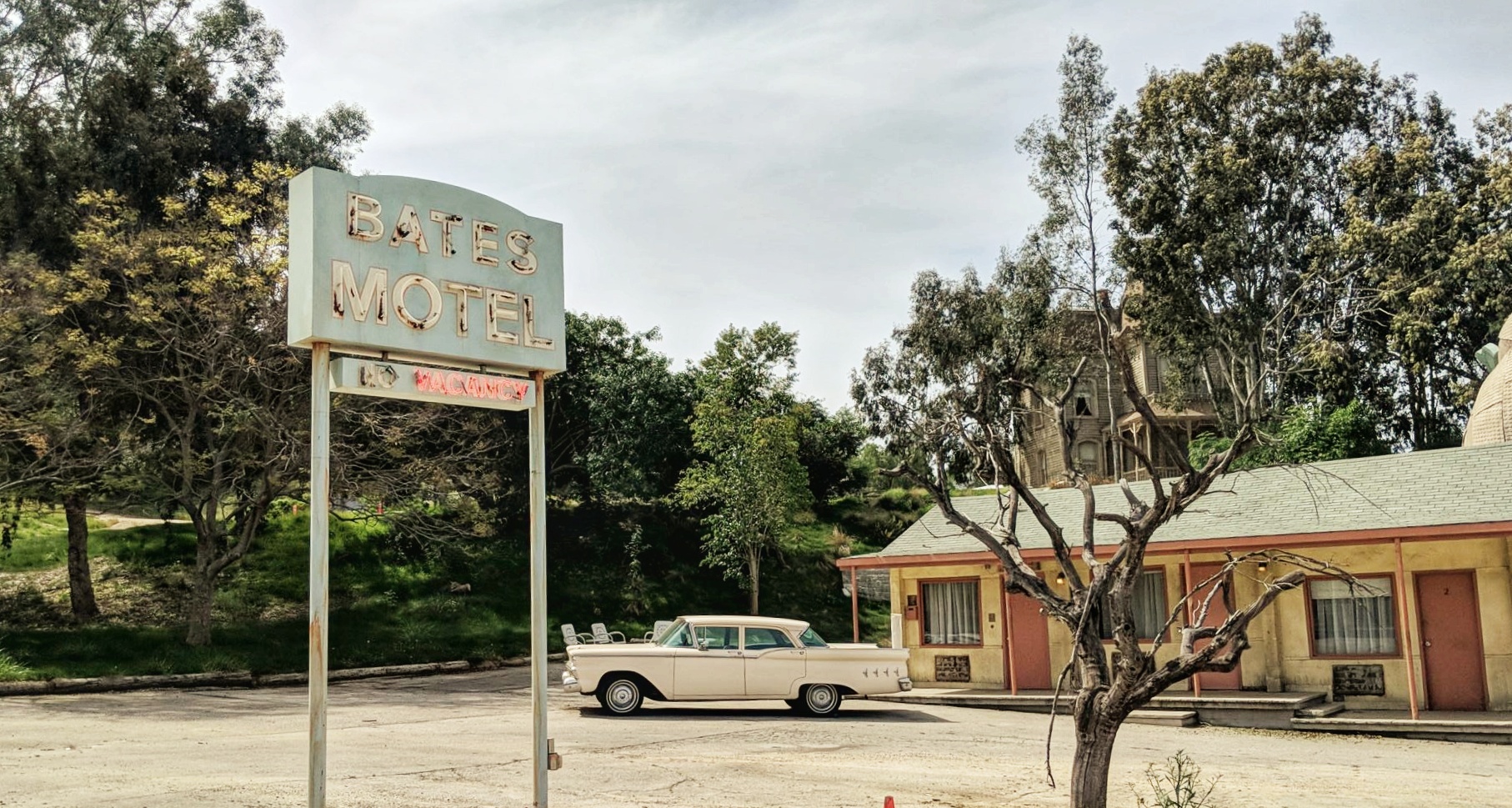 The Bates Motel set at Universal Studios Hollywood. There is a faded blue sign that reads Bates Motel in the foreground, and behind it you can see a classic car and an old motel set. The motel is painted in faded yellow, red and blue colours.