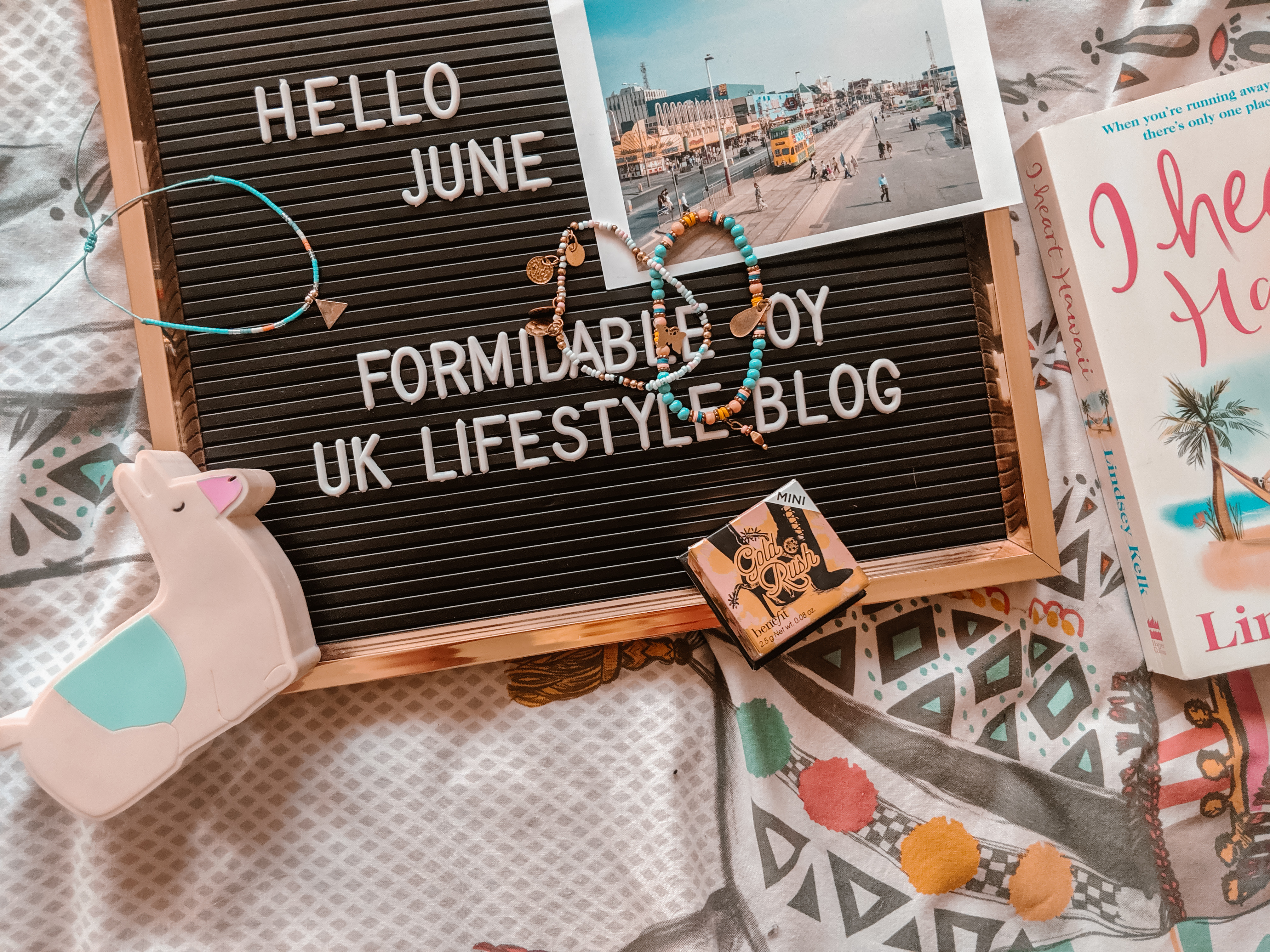 A flat lay image featuring a letter board displaying "HELLO JUNE FORMIDABLE JOY UK LIFESTYLE BLOG". Surrounding the board are two photos, a book, two bracelets, a necklace, a llama-shaped container, and a small box with ornate design, all placed on a patterned surface.
