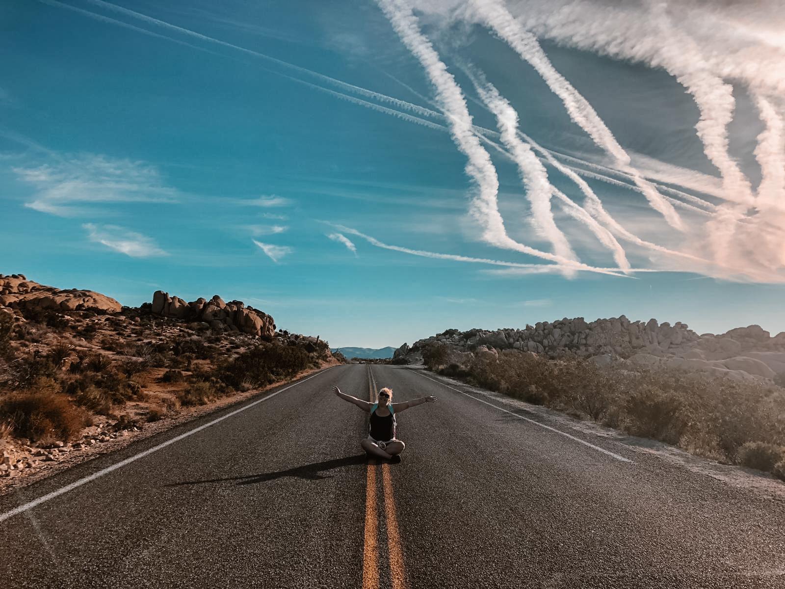 Louise is sitting down in the middle of a deserted road in America. There are is desert either side of her and a blue sky above. It's a traditional 'American road trip' photo.