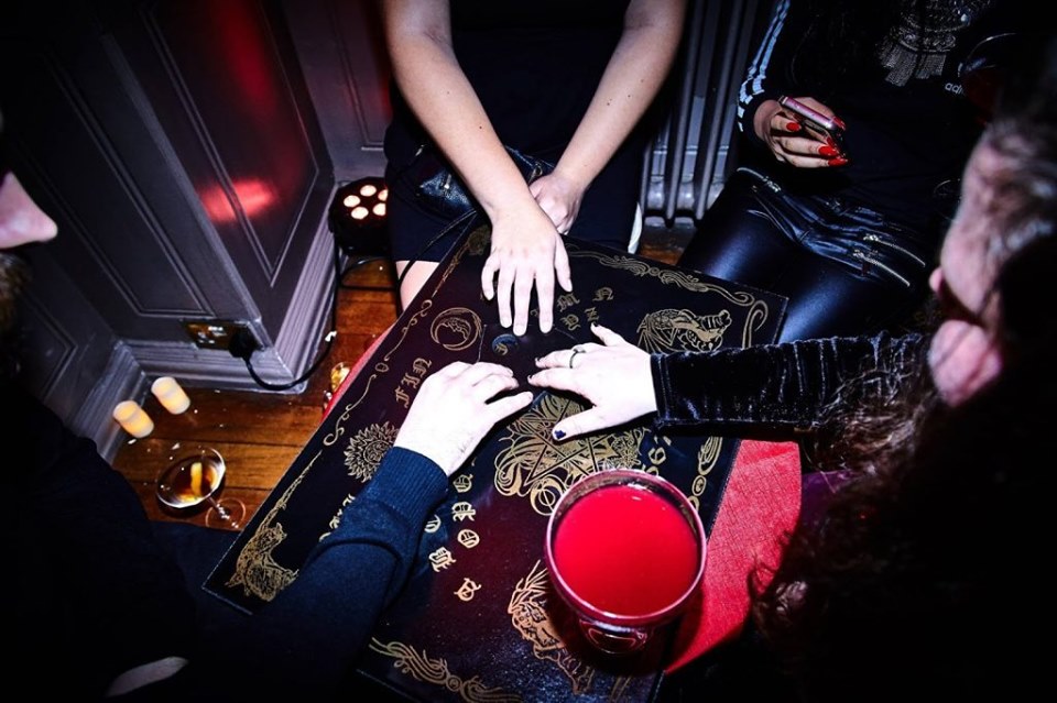 Louise and a crowd of people sat around a ouija board. The decor around them is dark and gothtic, with burning candles and deep red cocktails.