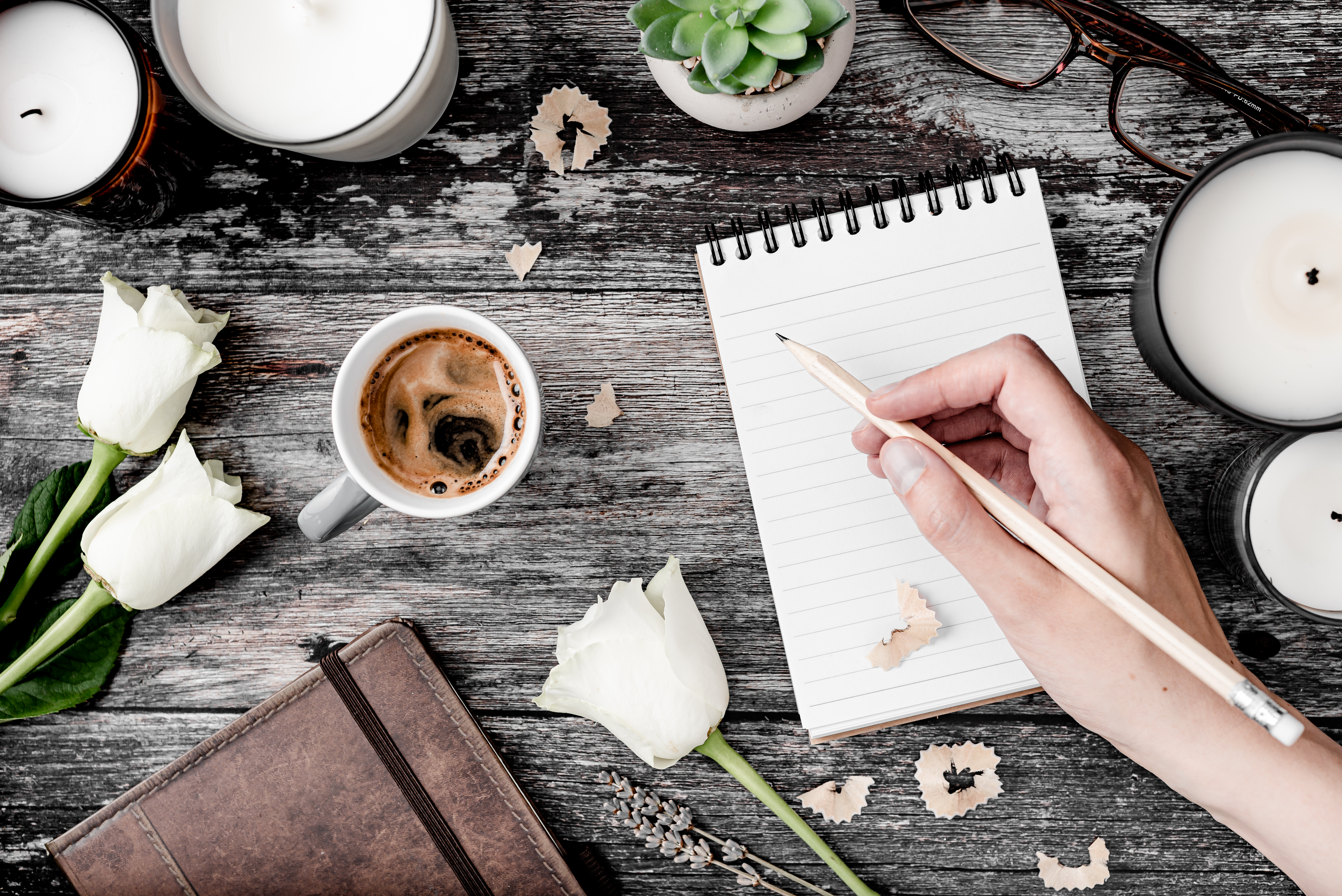 Hand Holding PenciL Over Notepad On Grey Wooden Table With Brown Leather Journal And Candles And Flowers by Tide And Tree