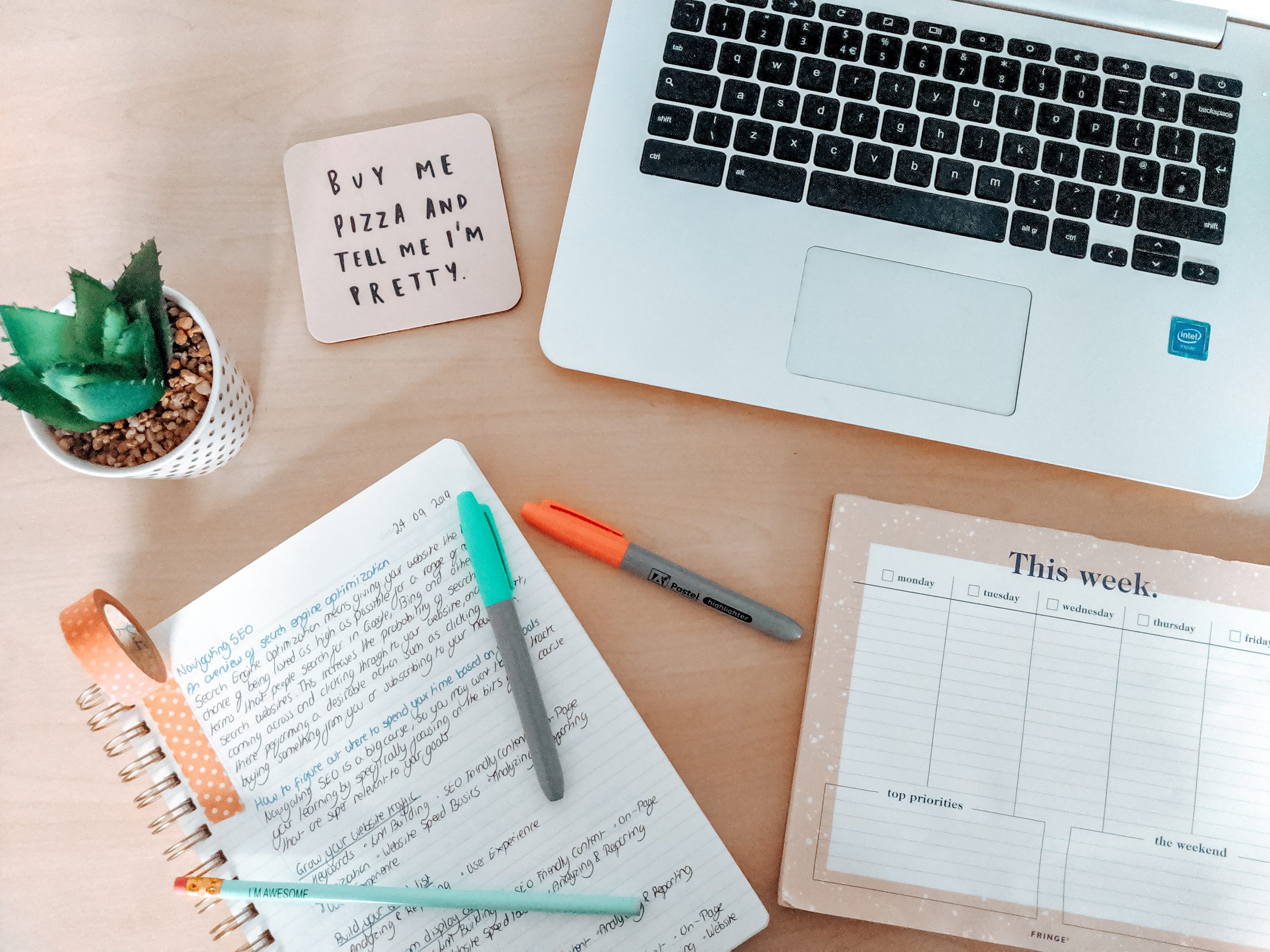 A desk with a laptop, a planner, a notebook with notes, pens, a potted succulent, and a coaster with the text "BUY ME PIZZA AND TELL ME I'M PRETTY!" sits alongside a weekly planner pad. The neat and organized scene suggests productivity in navigating SEO and effective planning.
