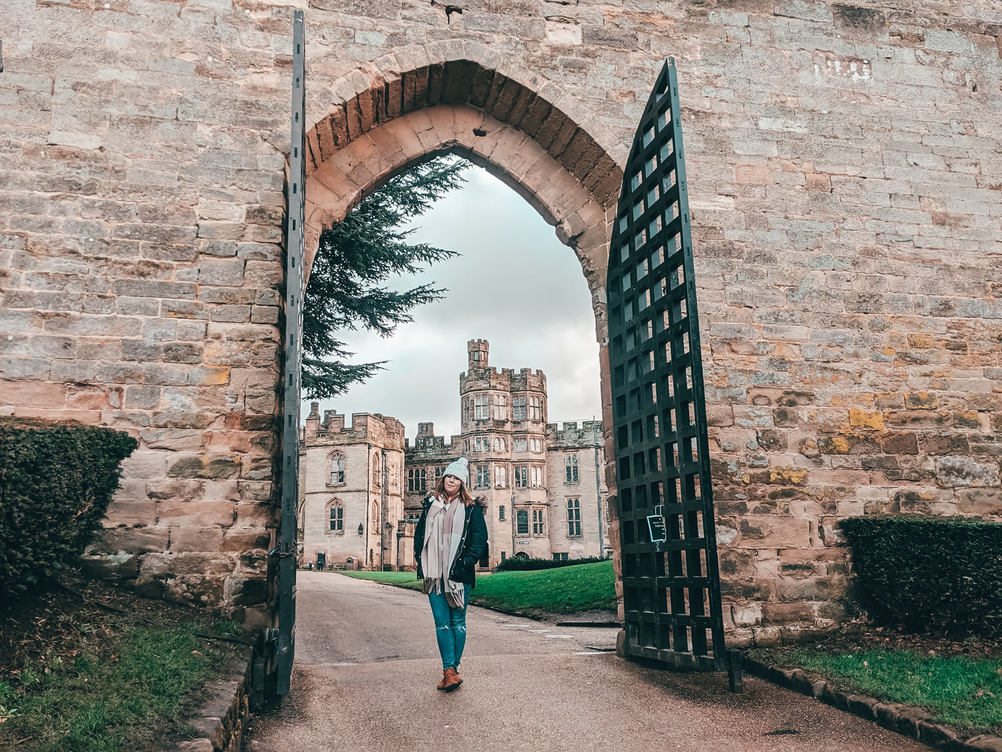 Louise walks through an open ancient stone archway with a partially open black metal gate, leading to Warwick Castle surrounded by greenery under a cloudy sky.