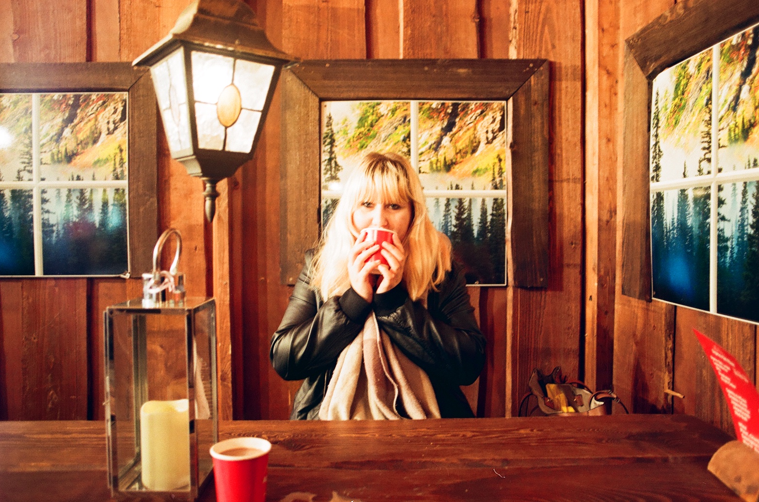 Louise with blonde hair is sitting in a cosy wooden room, sipping a hot beverage from a red cup. The room is warmly lit, with a lantern and framed landscape pictures on the walls behind them, embodying a seasonal nostalgic vibe as if from a Christmas movie. Another red cup and a candle in a glass holder are on the table in front.