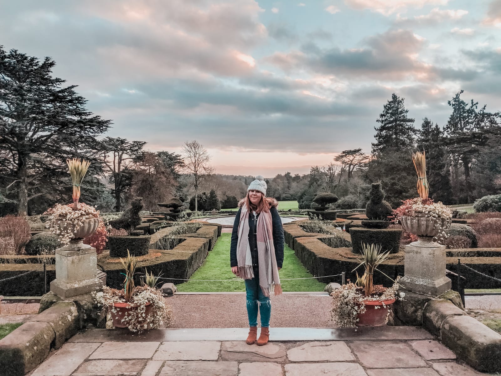 Louise stands on a patio in a formal garden during a partly cloudy 2020 sunset. They are wearing a coat, jeans, a scarf, and a knit hat. Behind them stretches a manicured garden with geometric hedges, potted plants, and a backdrop of trees and a distant hill.