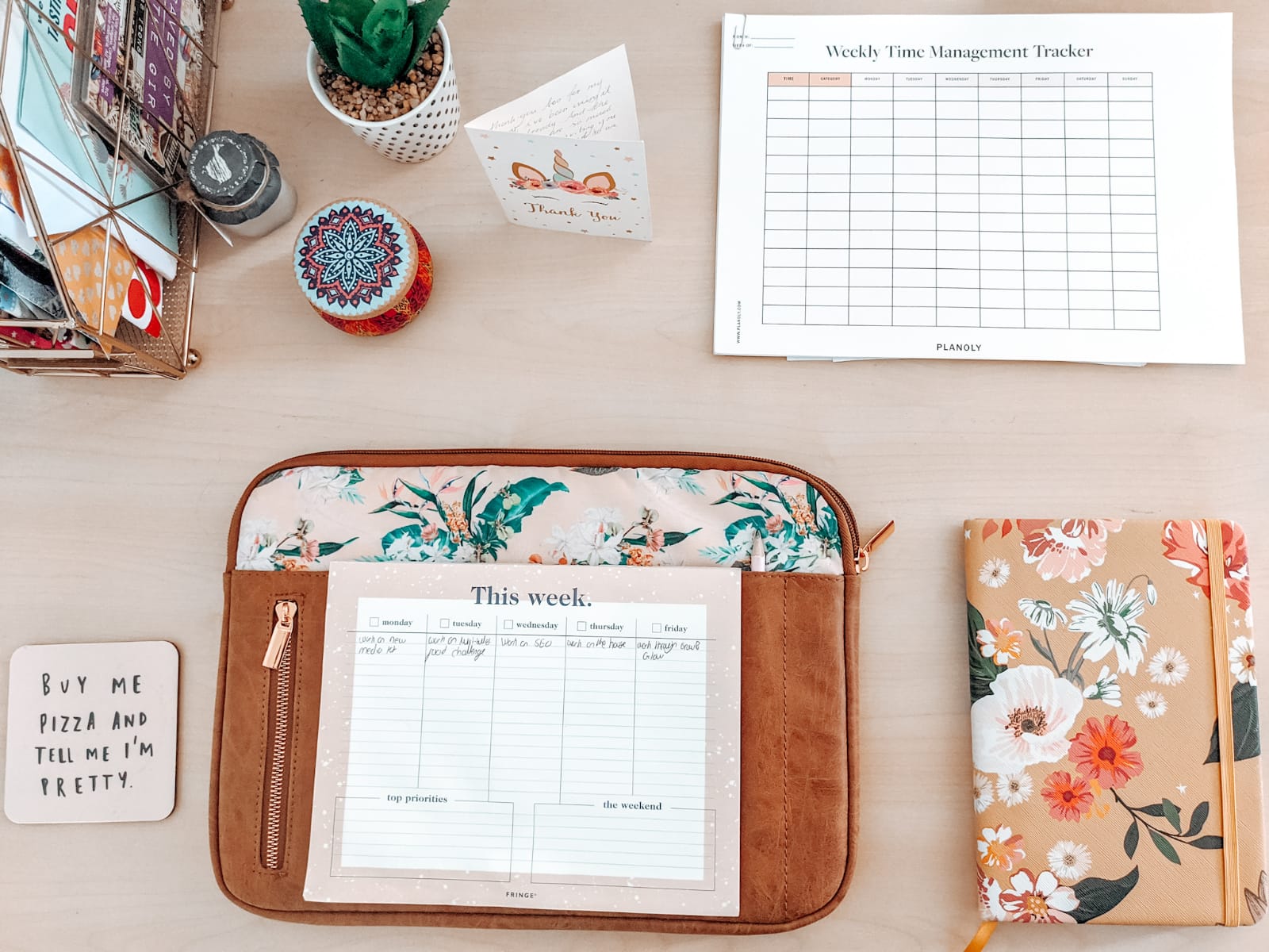 A neatly organized desk featuring a weekly planner, a time management tracker, stationary items, decorative objects, and houseplants, conveying a productive and aesthetic workspace for working from home.