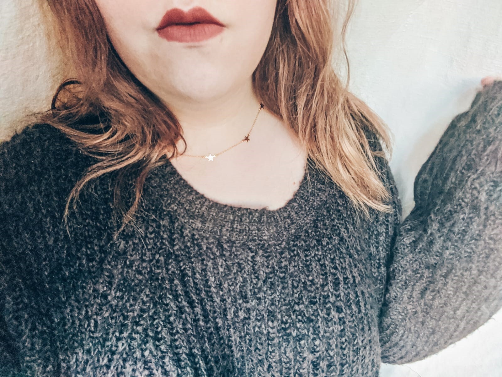 Louise with wavy hair is pictured from the chest up, wearing a dark gray knitted sweater. They are also wearing a delicate star necklet with star-shaped charms. The background is plain and light-colored.