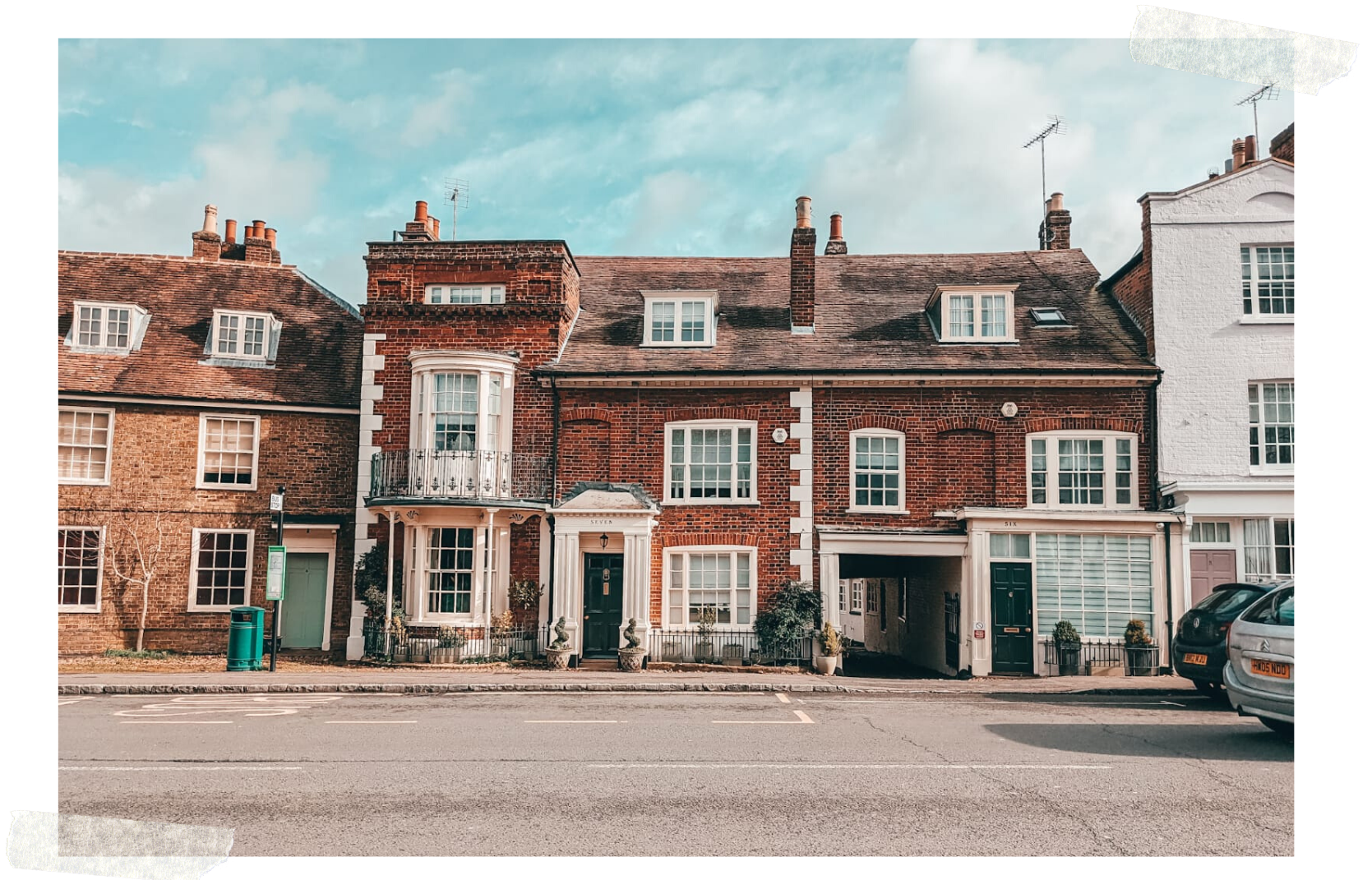 A quaint street lined with traditional red brick houses featuring classic British architecture, under a clear blue sky in February. A car is parked on the road in the foreground.