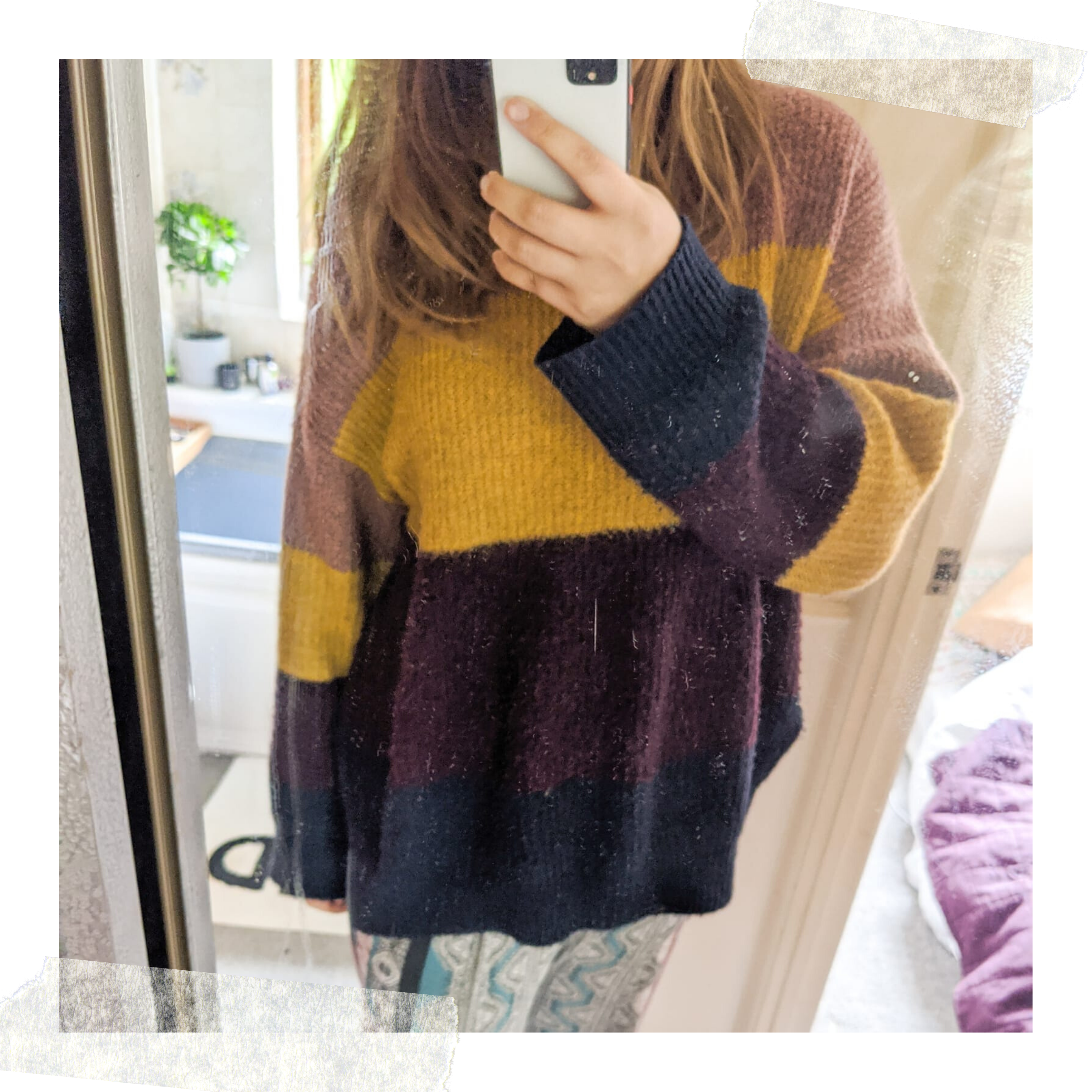 Louise stands in front of a mirror, taking a selfie. They are wearing a cozy, oversized sweater with wide horizontal stripes of yellow, burgundy, navy, and mauve—a typical day in lockdown attire. Their face is obscured by the phone, and the background shows a room with a bed and bathroom.