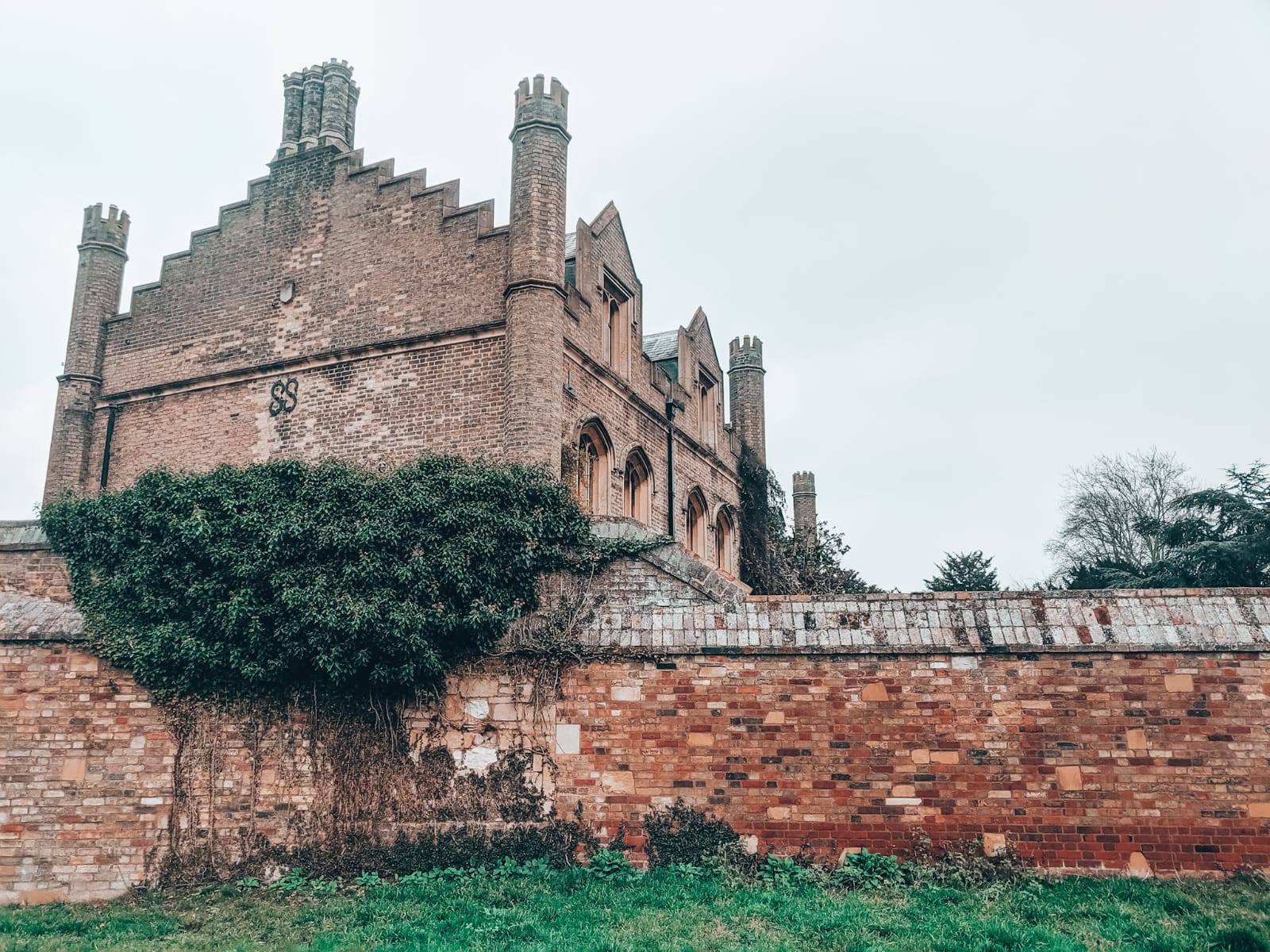 A historic red-brick building with a stepped gable and tall chimneys stands behind an old brick wall covered partially in ivy. Perfect for moody trips, the overcast sky above enhances the scene's atmosphere, while green grass spreads invitingly in the foreground.