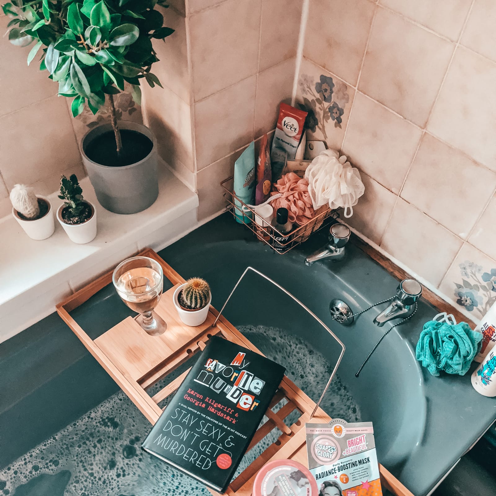 A bathtub scene with a wooden tray holding a book titled "Stay Sexy & Don't Get Murdered," a glass of white wine, and a potted cactus. The bathtub has bubbles, and nearby are various bathing items, including a plant, scrubbers, and a face mask packet.