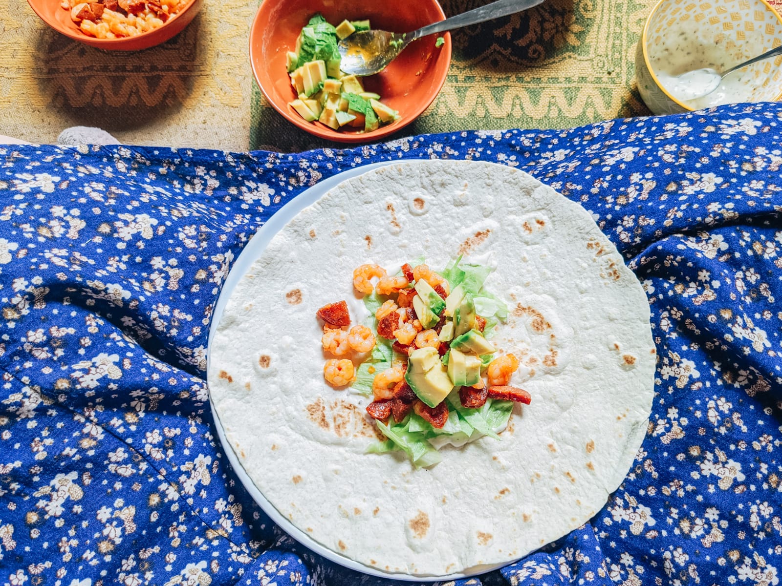 A close-up of a tortilla wrapped on a blue floral-patterned lap, recreating our favorite Gousto meals. The tortilla is topped with shrimp, avocado, lettuce, and other ingredients. Nearby, there are small bowls with additional toppings and a spoon.