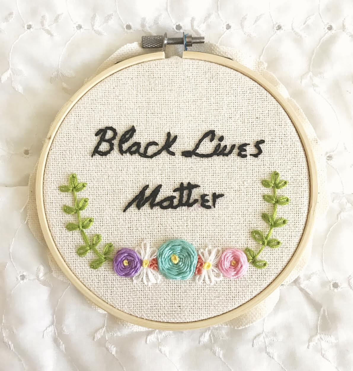 An embroidery hoop on a white lace fabric elegantly displays the words "Black Lives Matter" stitched in black thread. Below the powerful text, vibrant embroidered flowers and lush green leaves add a beautiful decorative touch.
