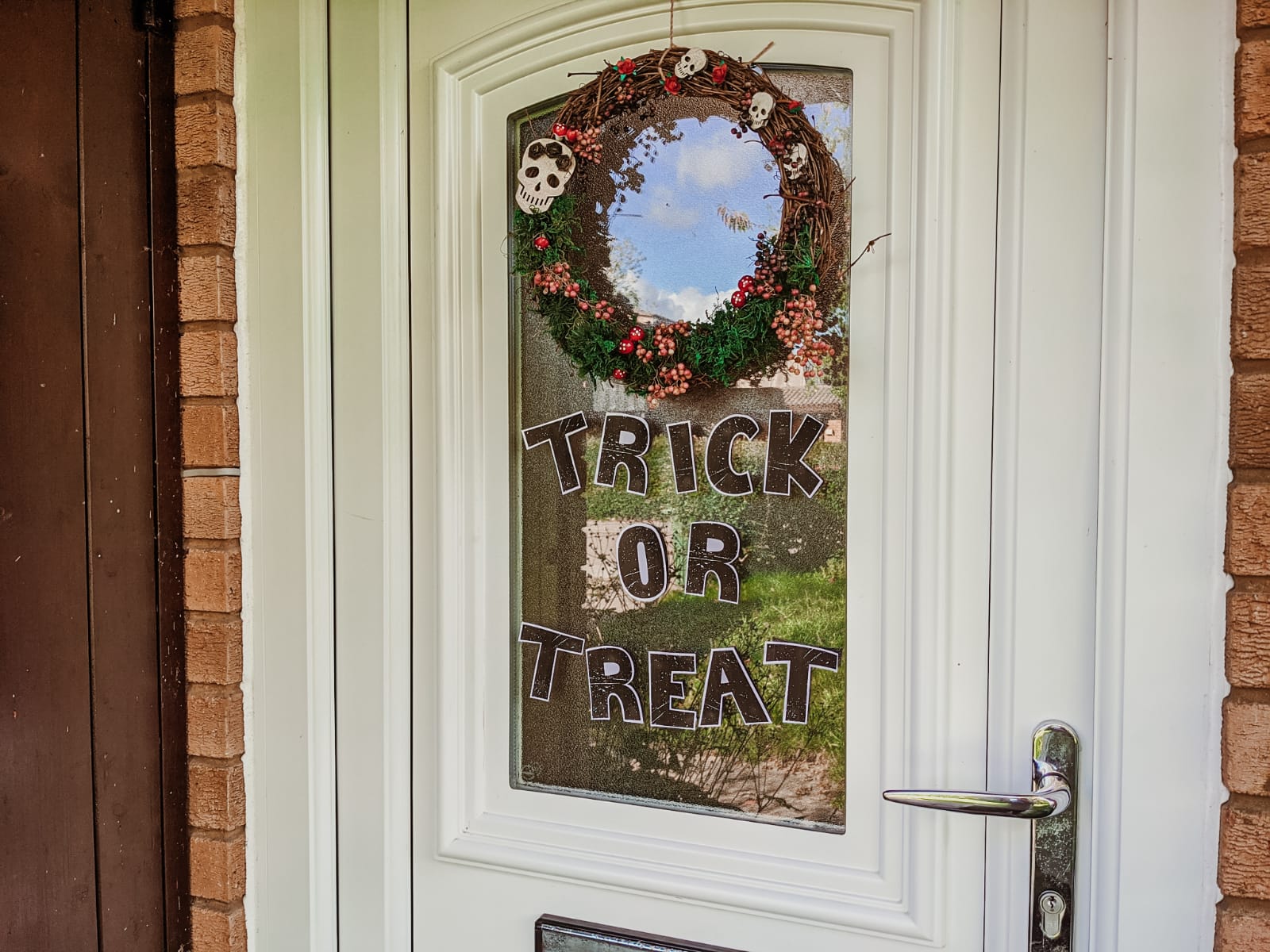 A white front door is decorated for Halloween with a wreath featuring red berries and small skull ornaments. On the door's glass pane, the words "TRICK OR TREAT" are displayed in black cut out letters.