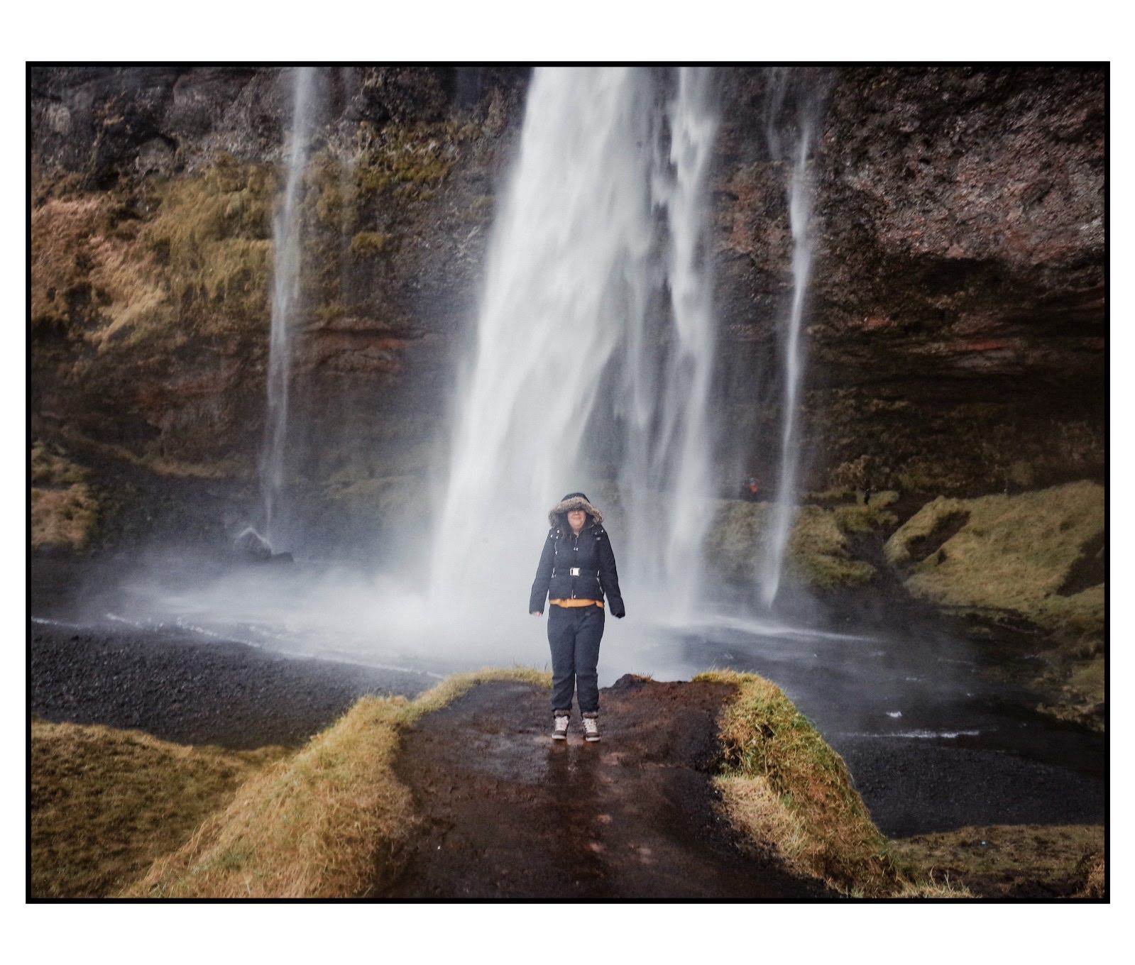 Louise stands on a pathway in Southern Iceland, facing a majestic waterfall, wearing a dark jacket and backpack, mist and spray surrounding them in a lush, rocky landscape. It's the perfect photo to represent my Iceland Travel Diary.