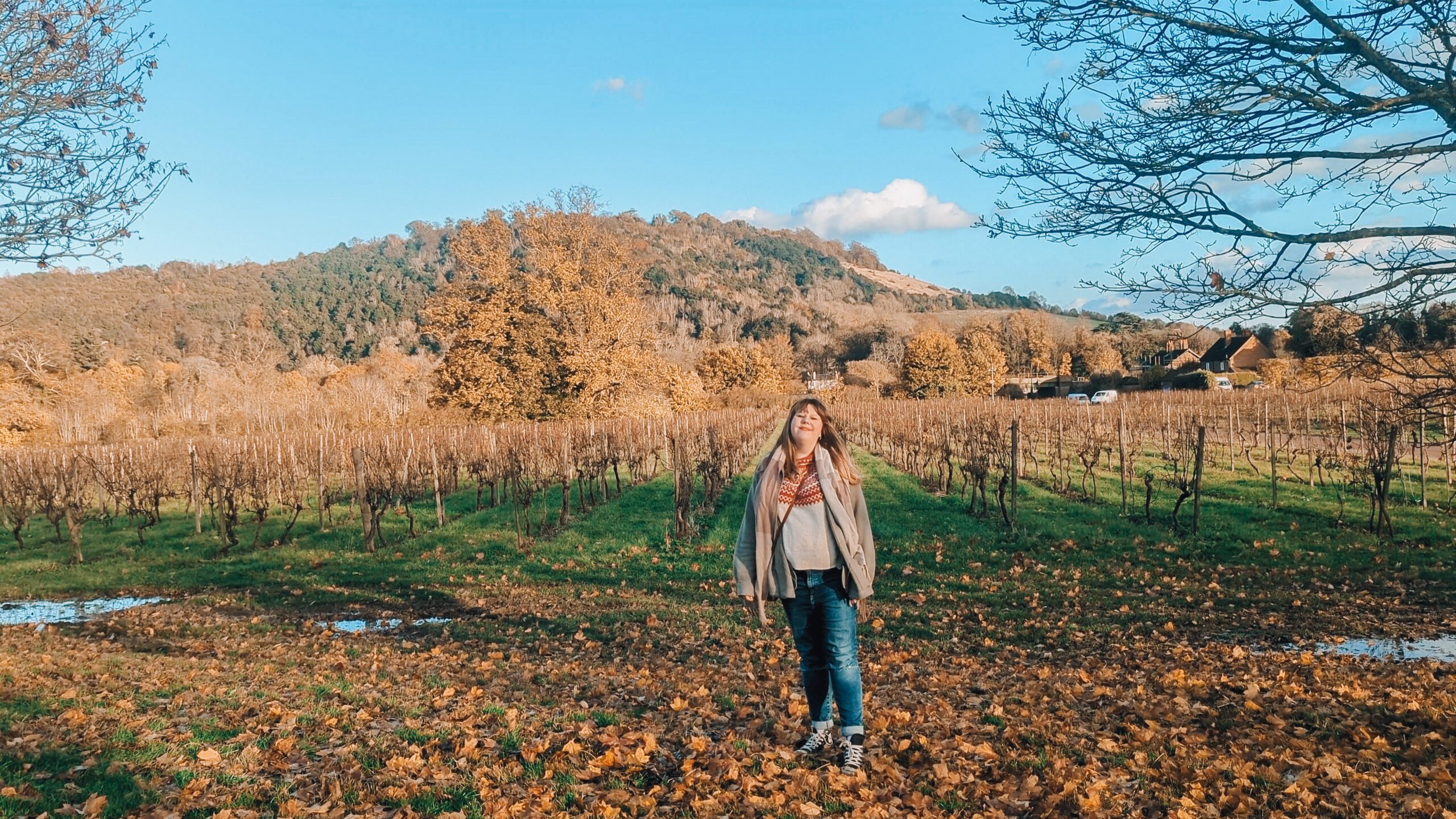 Louise stands on Denbies Wine Estate, with the rolling hills of Surrey behind her. Bright orange leaves have fallen to the ground and the trees on the hills are in rich shades of orange, despite it being winter.